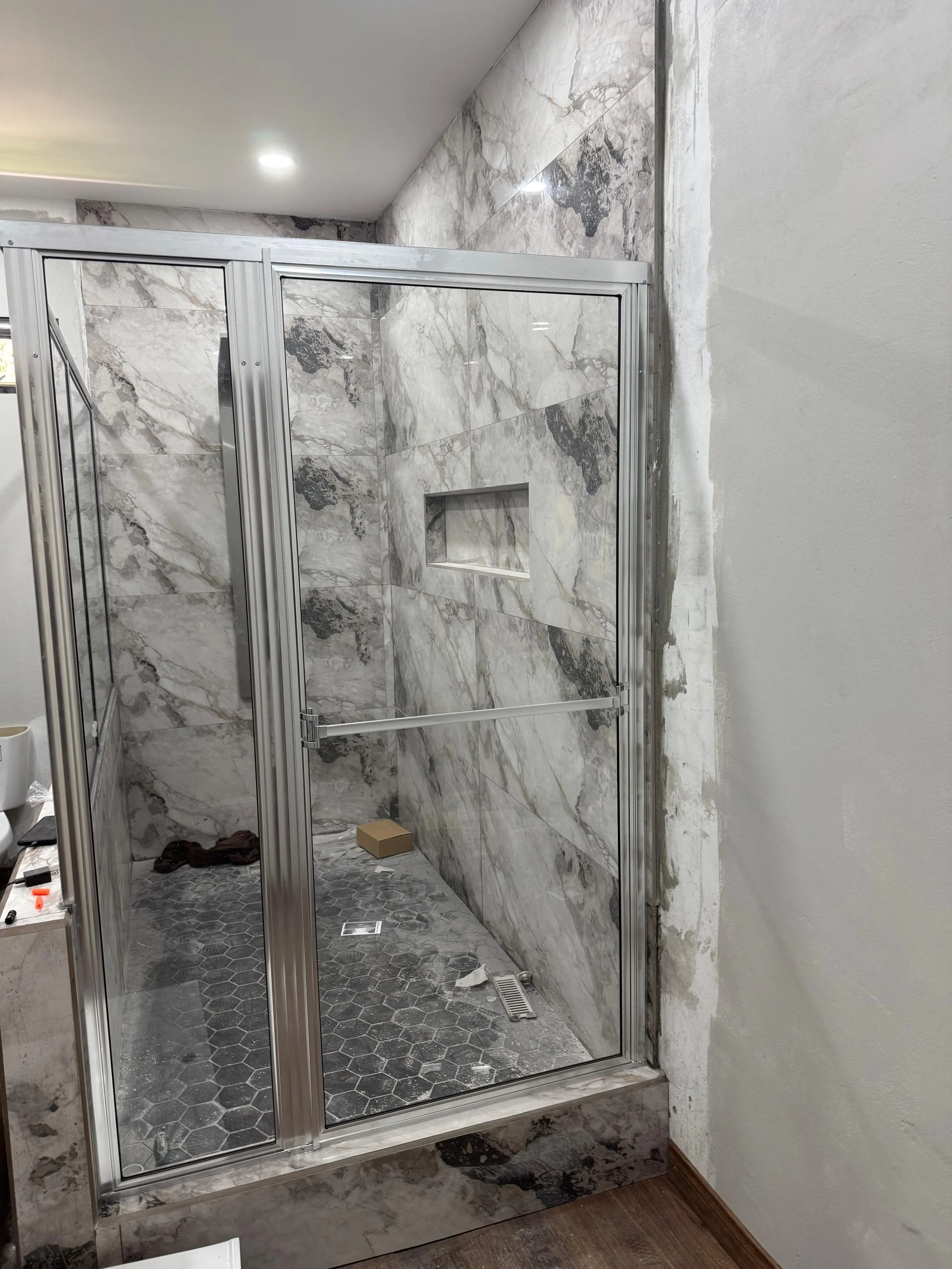 Modern shower with marble walls, glass doors, hexagonal floor tiles, and a built-in shelf, under construction.