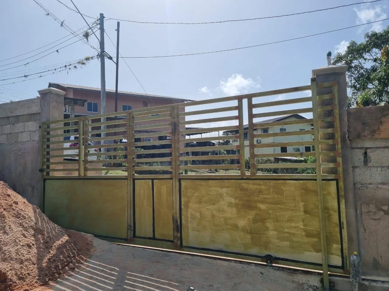 Metal gate with wooden panels, mounted on concrete posts, situated in front of a residential area with houses and power lines in the background.