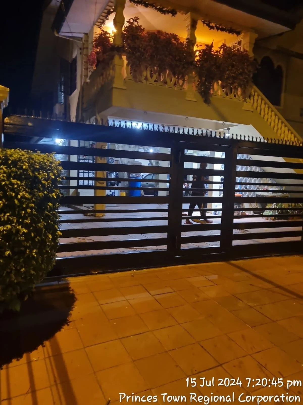 Nighttime scene of a house with a porch, second-floor balcony, and stairs with decorative railings, surrounded by trees and plants. The area is illuminated by outdoor lights. A person is standing near the gate, which is made of horizontal bars. The g