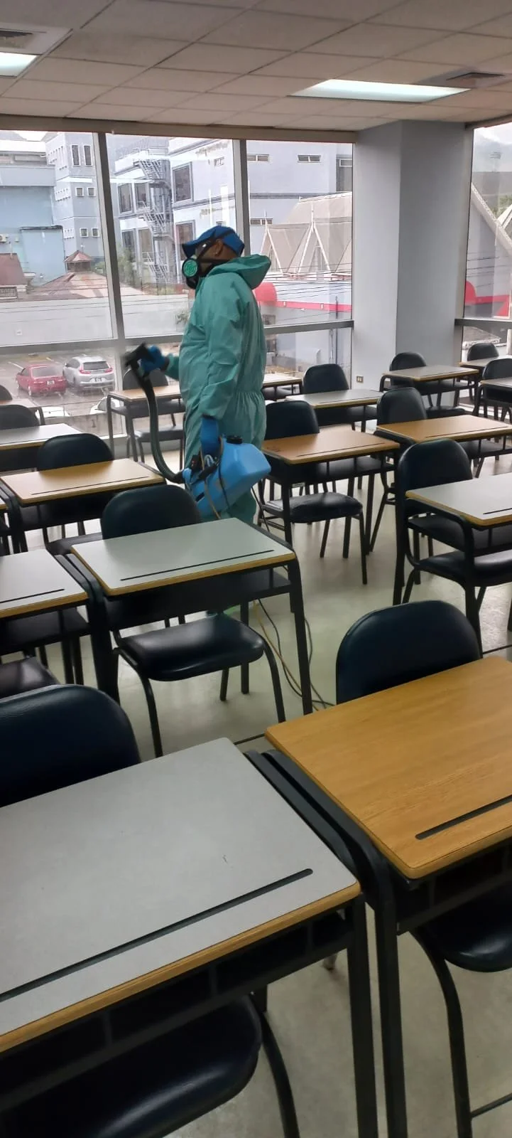 A person in protective gear disinfecting a classroom with empty desks and chairs, large windows showing an urban area outside.