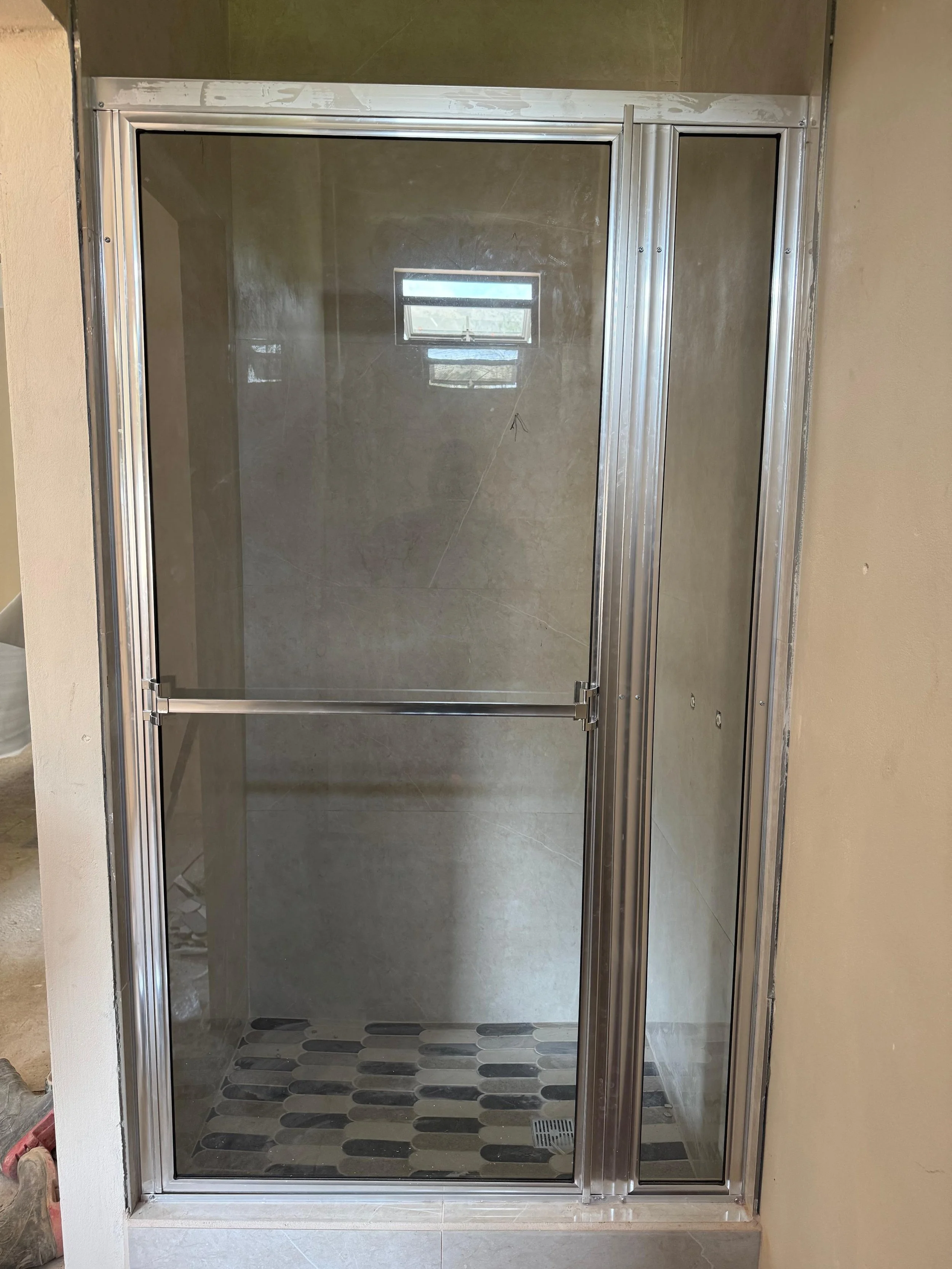 A glass shower stall with metal trim, installed in a bathroom with beige and green walls, and a small window visible through the glass.