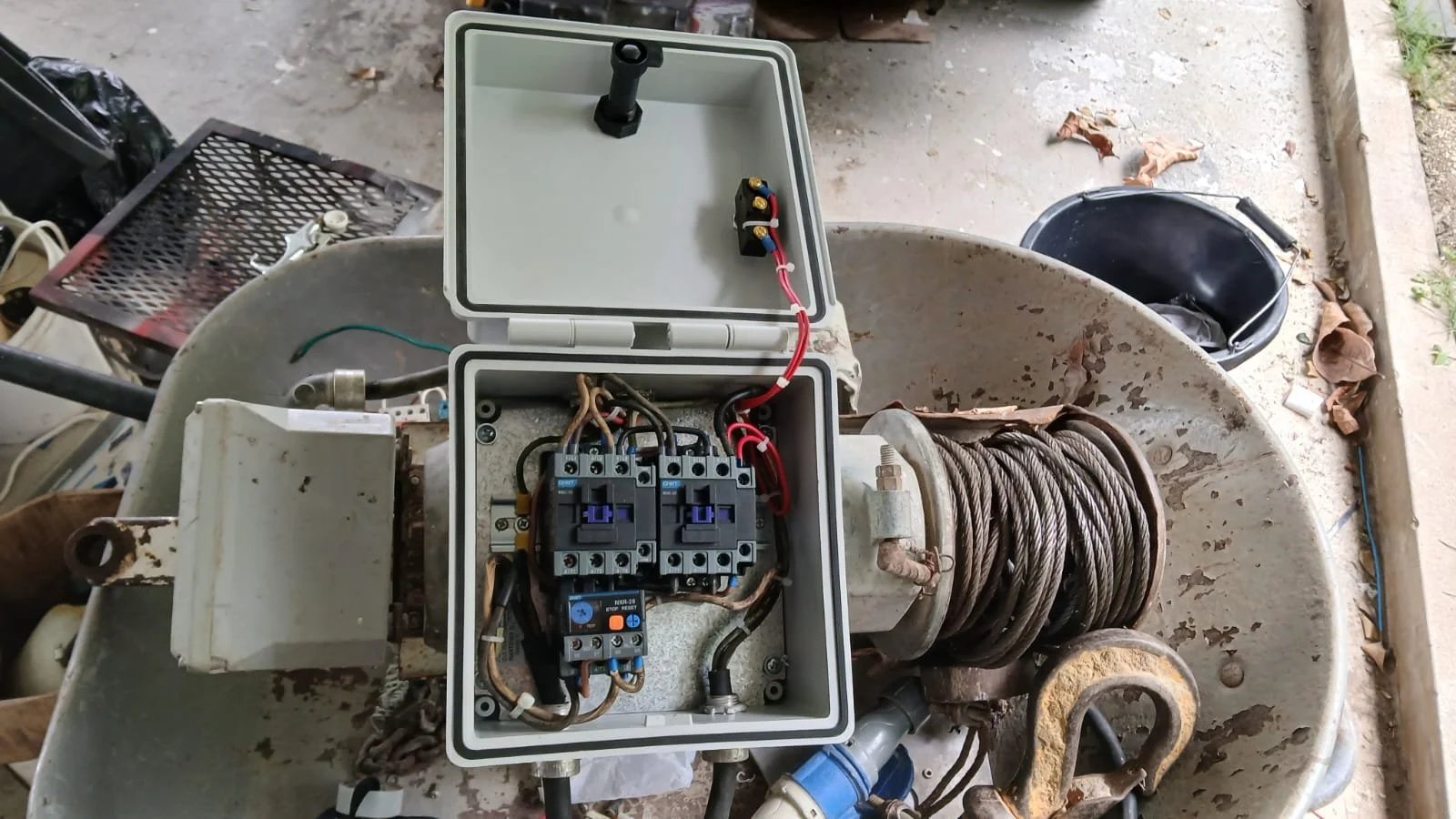 Close-up view of an electrical control box with wiring and circuit breakers, mounted on an old, rusted cable reel, outdoors on a concrete surface.
