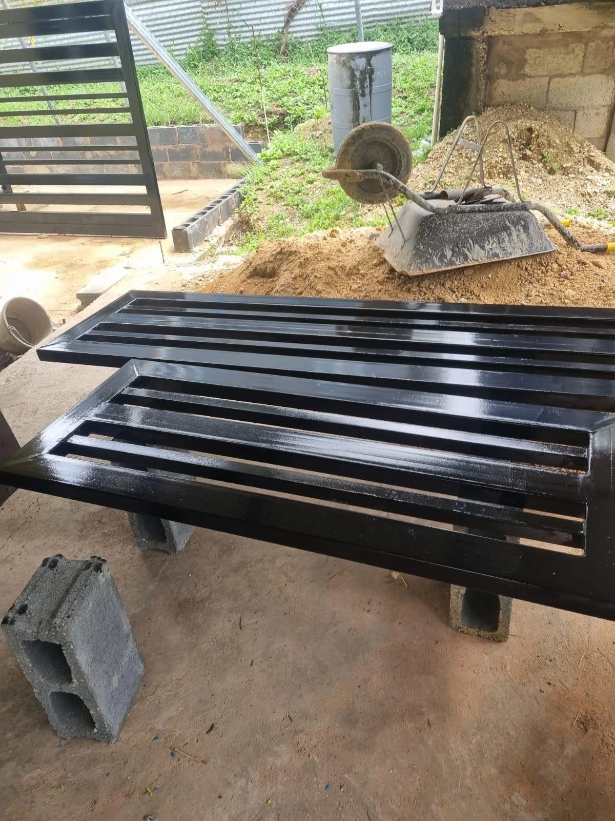 Two black metal grill panels resting on cinder blocks in a partially built outdoor area. In the background, there is a pile of dirt, a shovel, a discarded wheelbarrow, and a metal barrel near a brick and concrete wall.