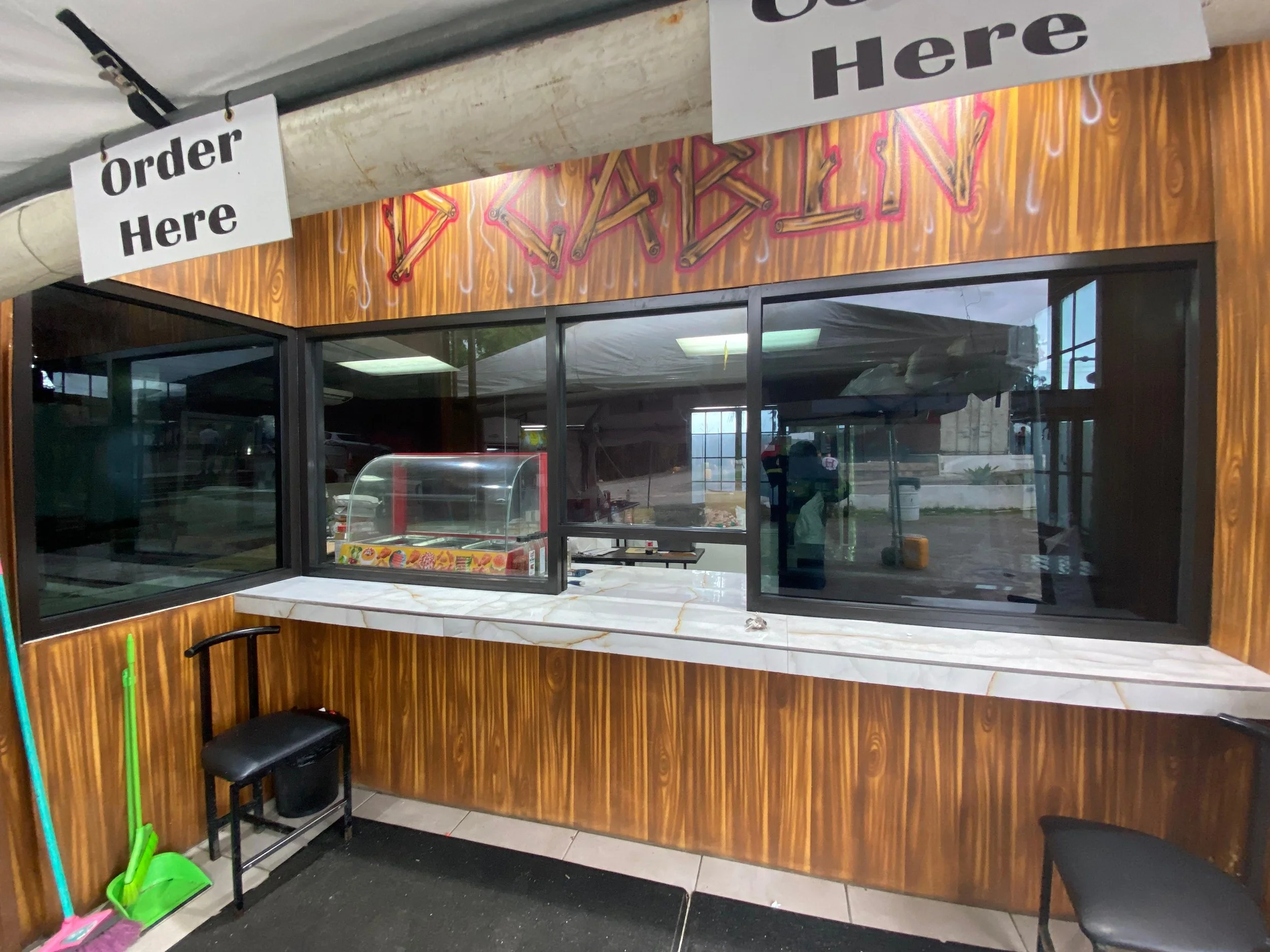 View of a fast-food restaurant window counter with signs that say 'Order Here' and 'Here', a small stool, cleaning tools, and a display case inside. Outside, reflections show a parking lot and cloudy sky.
