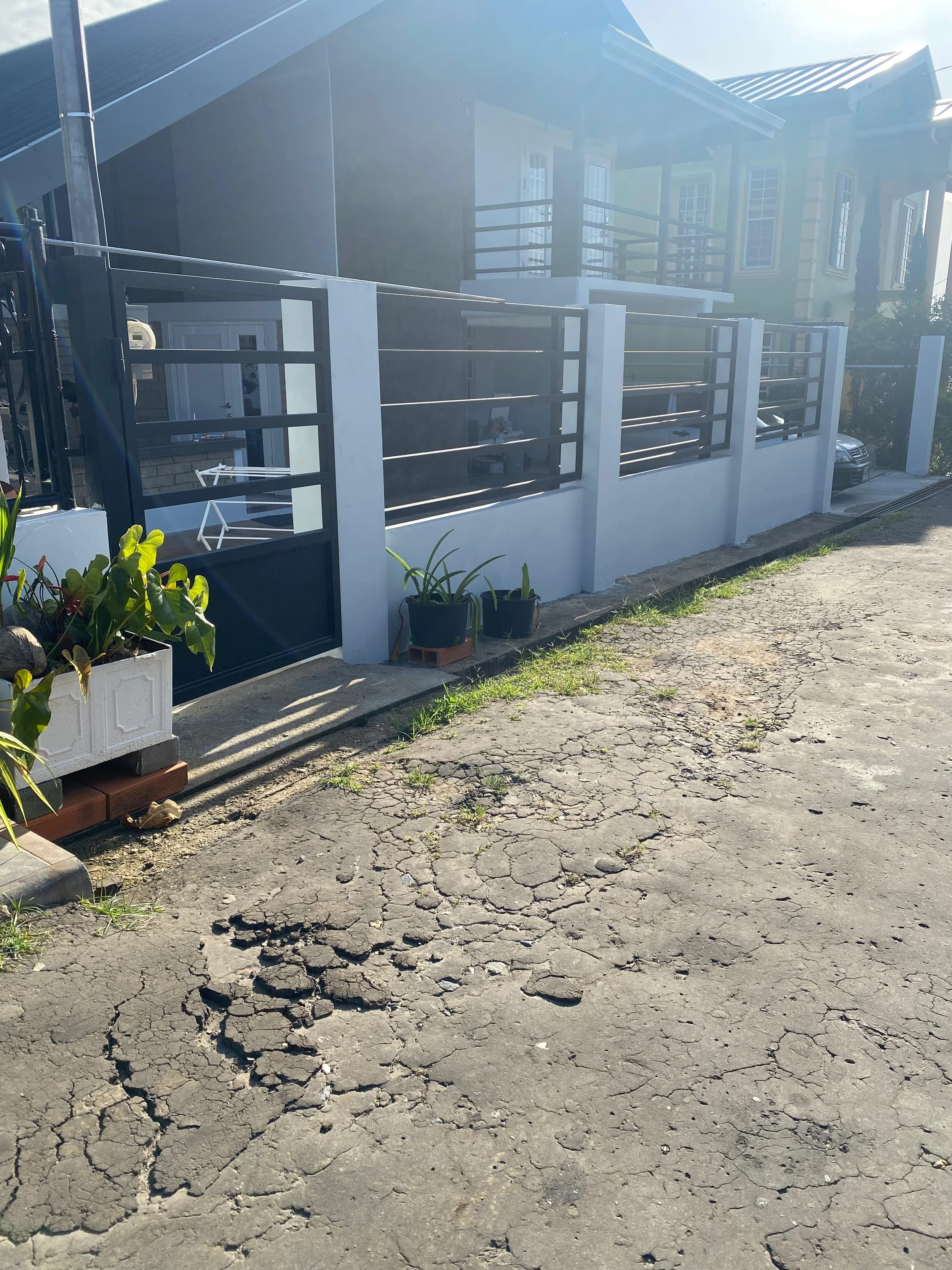 A residential house with a courtyard, black metal fencing, potted plants, and a parked car on a cracked asphalt driveway, seen in bright sunlight.