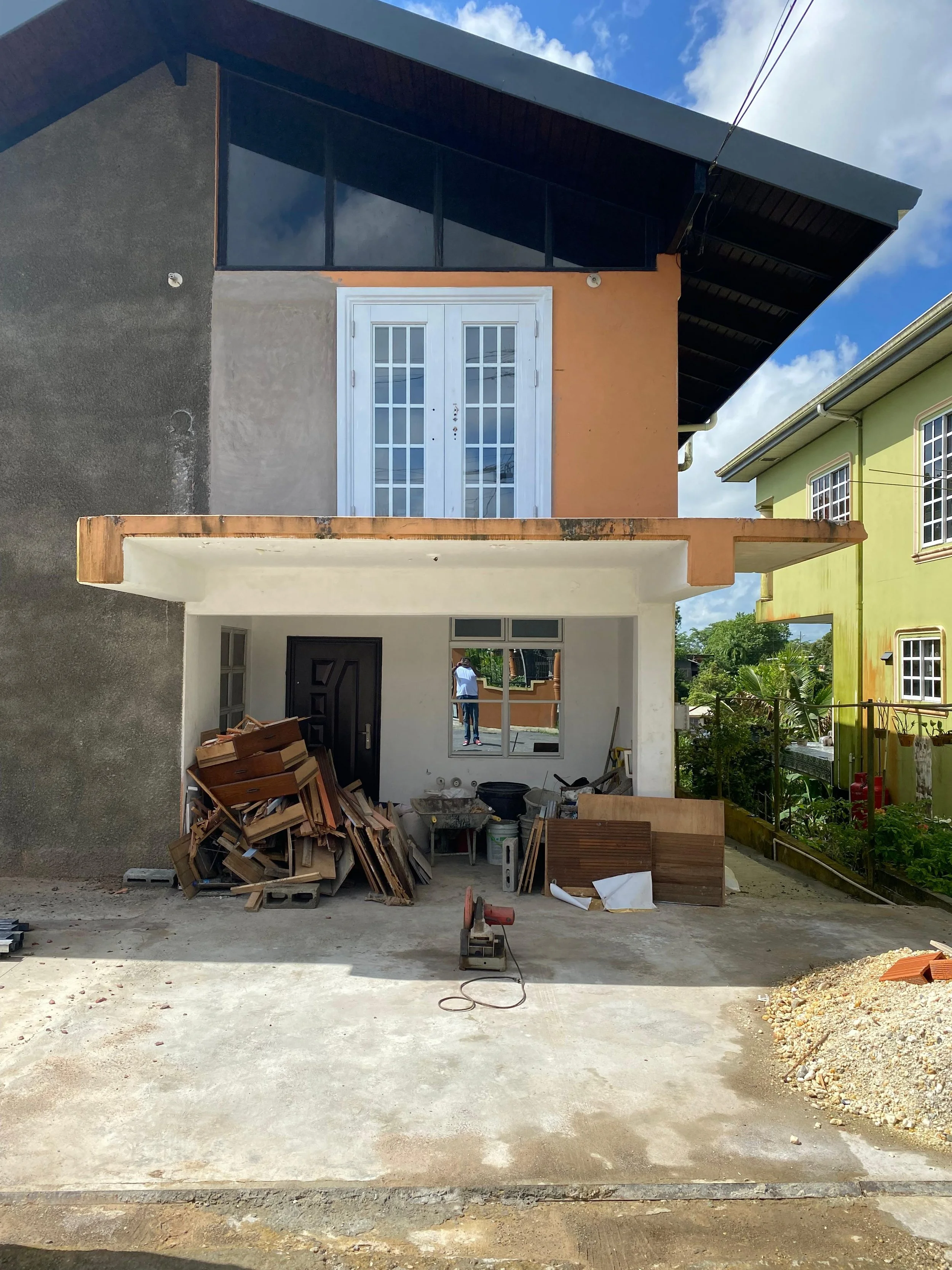 Construction site showing a partially built house with a covered garage, construction materials, and tools in the foreground, and multi-colored houses in the background.