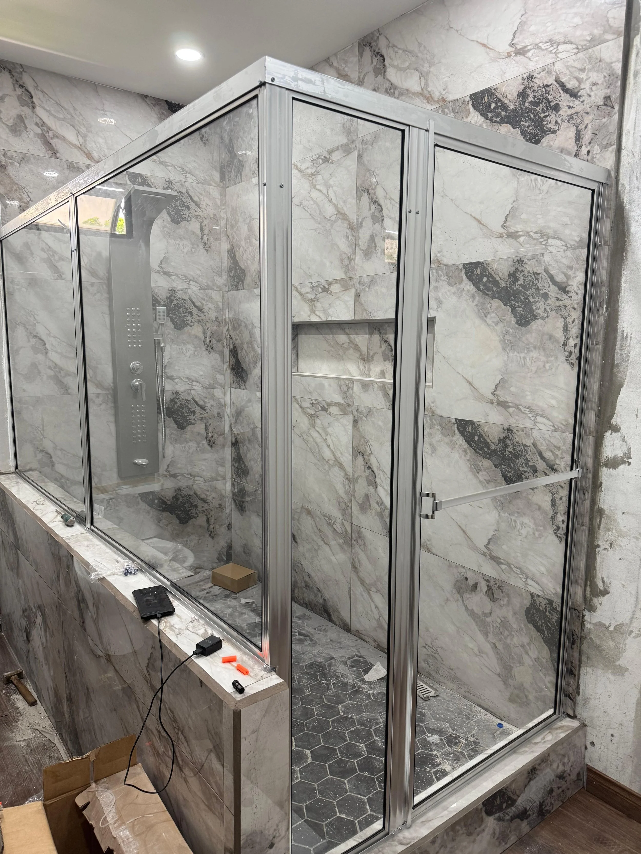 A marble finished shower enclosure with metal frames, glass doors, and a built-in shelf, situated in a marble bathroom with hexagonal tile flooring.