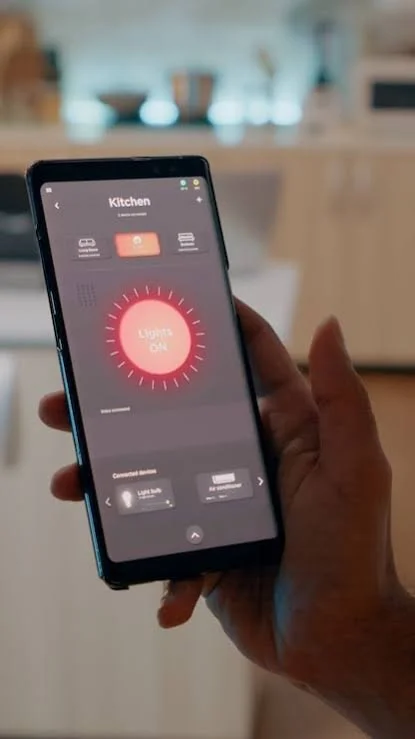Person holding a smartphone with a smart home app open, controlling kitchen lights that are turned on.