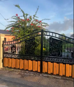 A black metal fence with decorative scrollwork, supported by a wooden base with vertical slats, set in a residential area with trees and a house in the background under a partly cloudy sky.