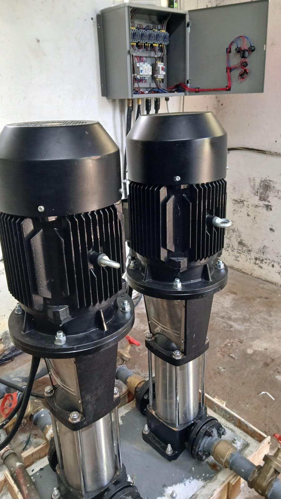 Two vertical water pumps with electric motors, pipes, and an electrical control box mounted on the wall.