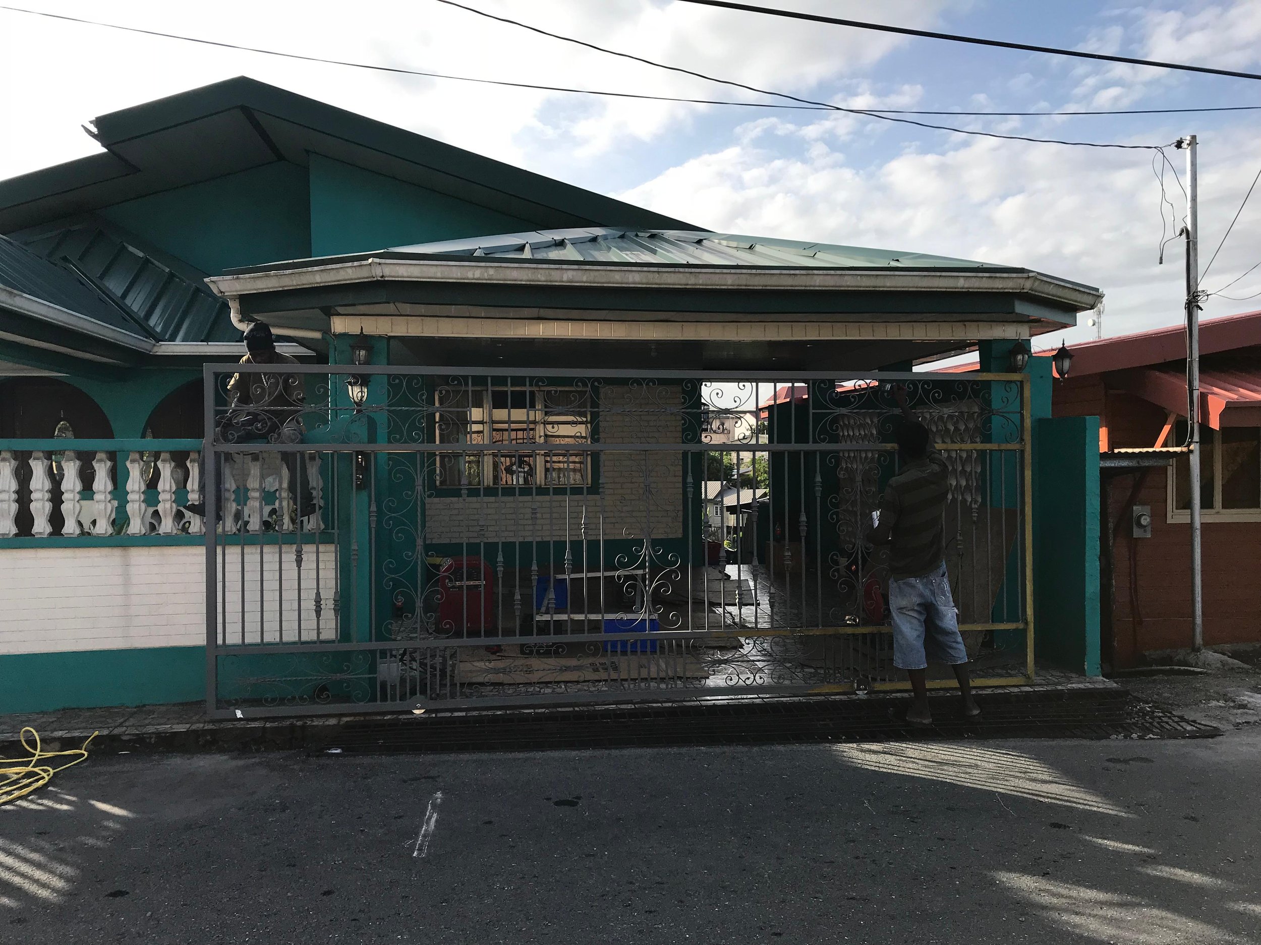 A turquoise house with a grey roof, iron gate, and a man in a striped shirt and shorts standing outside near the gate. Two people are on the roof working or inspecting it. Overhead power lines are in the sky.