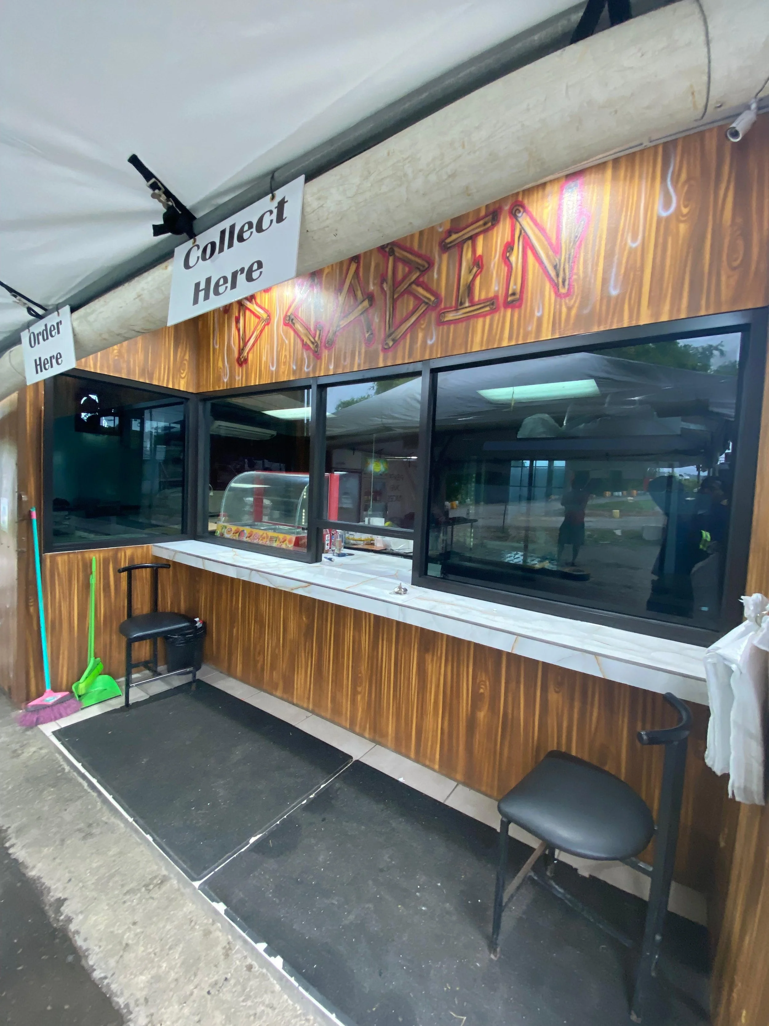 Food stand with a neon sign that spells out 'CRABIN', windows, and a small service counter, with cleaning supplies and chairs outside.
