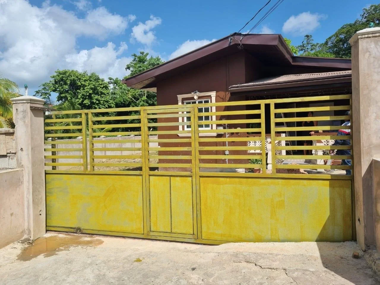 Yellow metal gate in front of a brown house with white framed window, surrounded by trees and a concrete sidewalk.