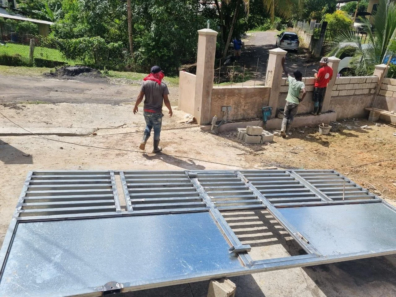 Construction workers installing a brick wall on a residential property, with metal framework and construction tools in the foreground and lush greenery in the background.
