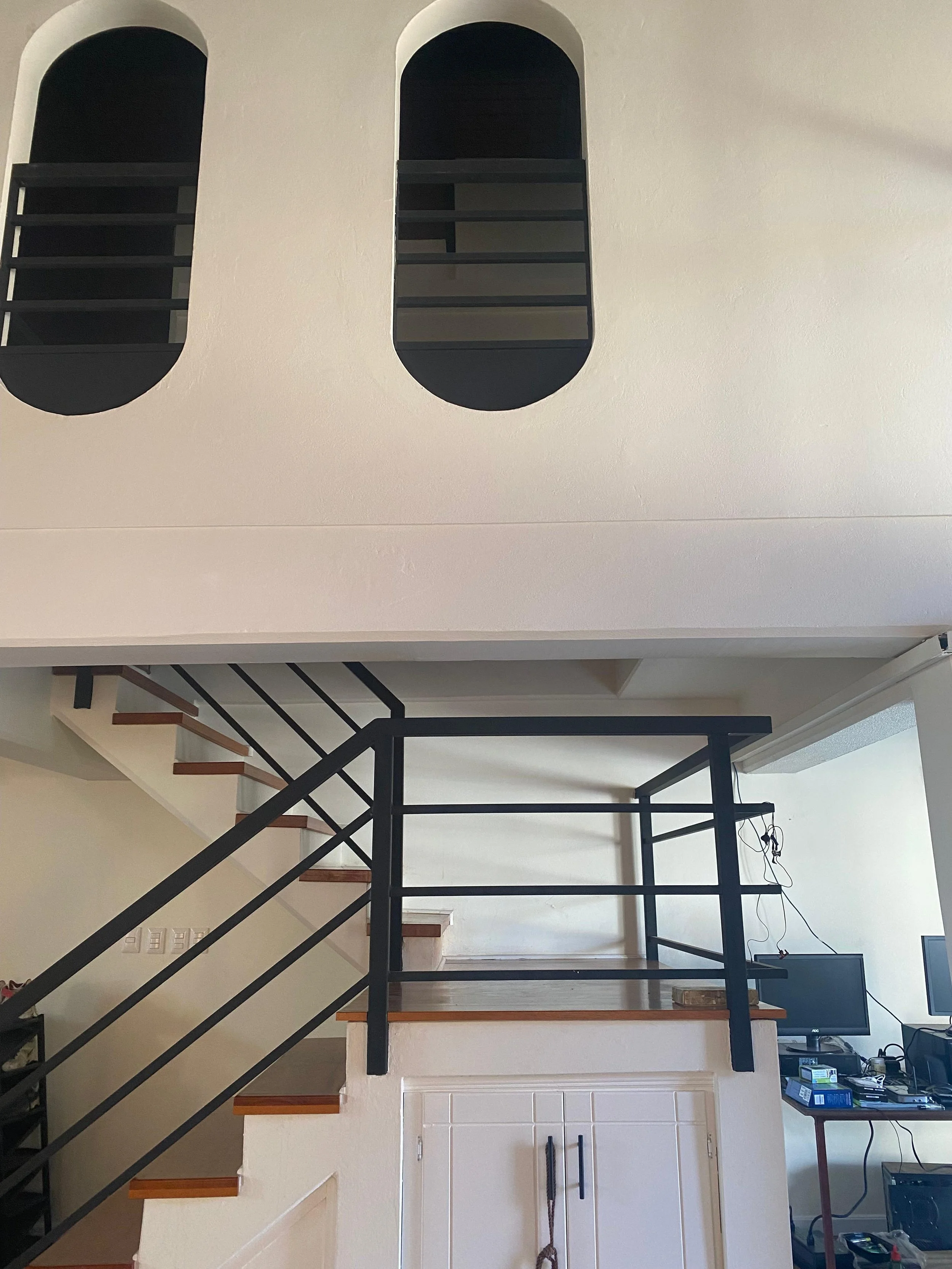 Image of a multi-level staircase area with black metal railings, wooden stairs, and a white wall. There is a flat-screen TV, a red-topped table with various electronic items, and a tangled cord hanging on the wall.