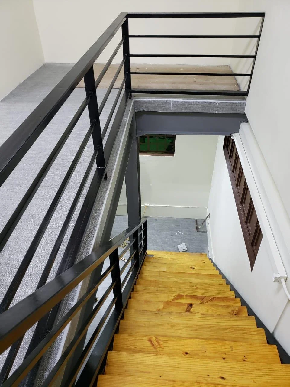 Interior view of a staircase with wooden steps and black metal railing, overlooking a lower floor with a grey carpeted floor and a small window.