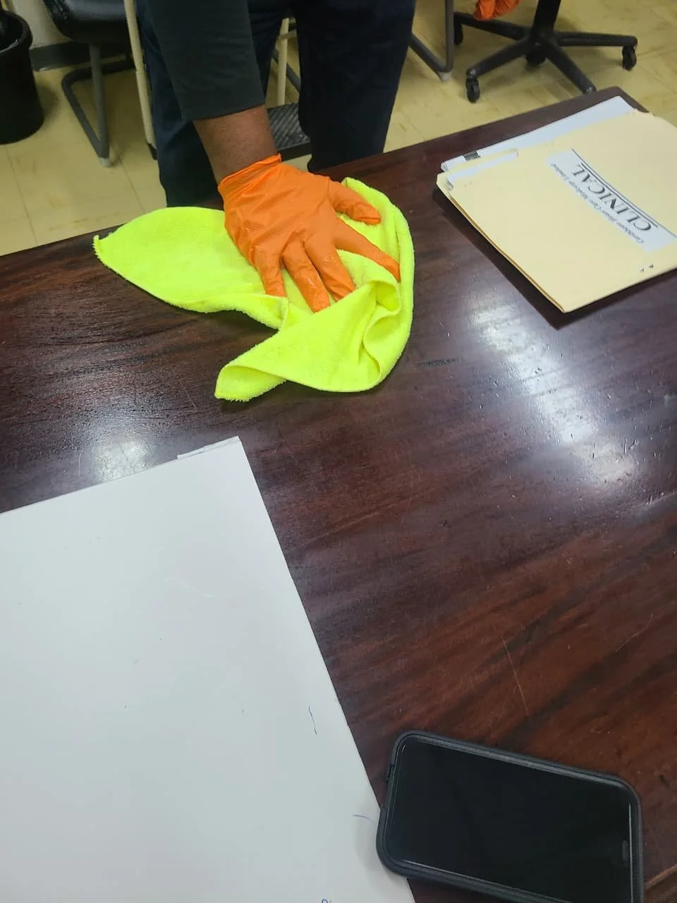 Person wearing an orange glove cleaning a dark wooden table with a yellow cloth. Papers, a clipboard, and a smartphone are on the table.