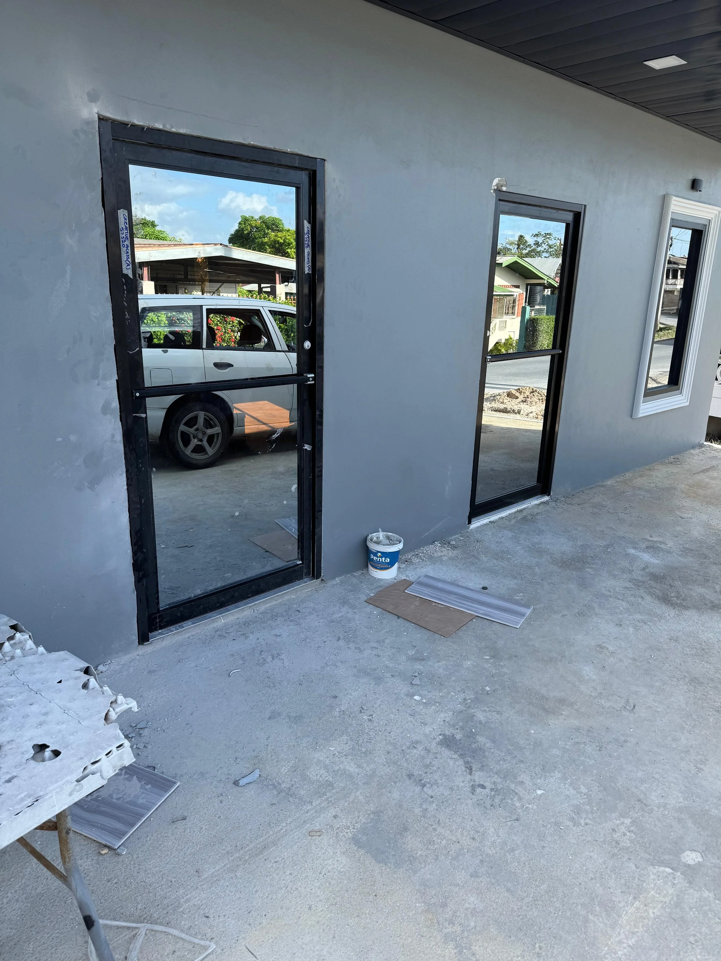 A construction site showing a gray wall with two window frames installed, one door frame installed, and construction tools and materials on the ground.