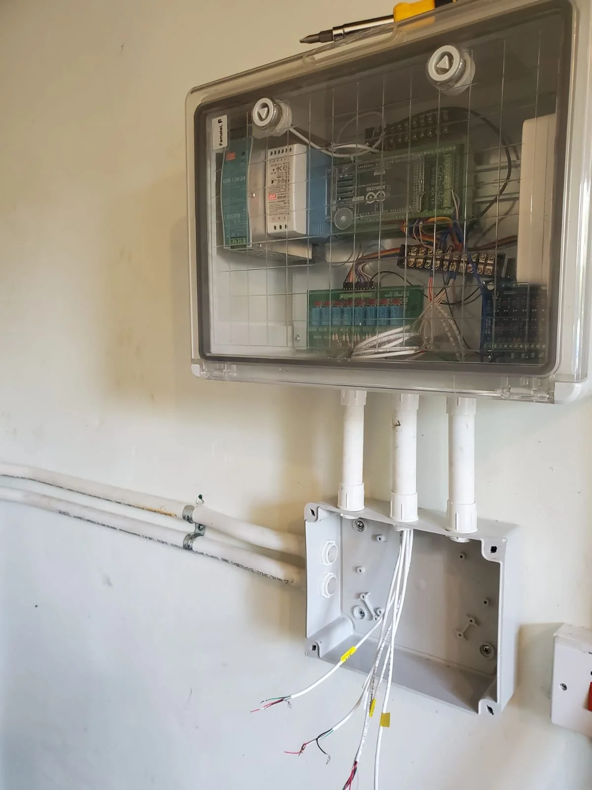 An electrical control box mounted on a wall with wires and components inside, and conduit pipes extending from the bottom. An empty wall-mounted box with wires protruding is below the control box.