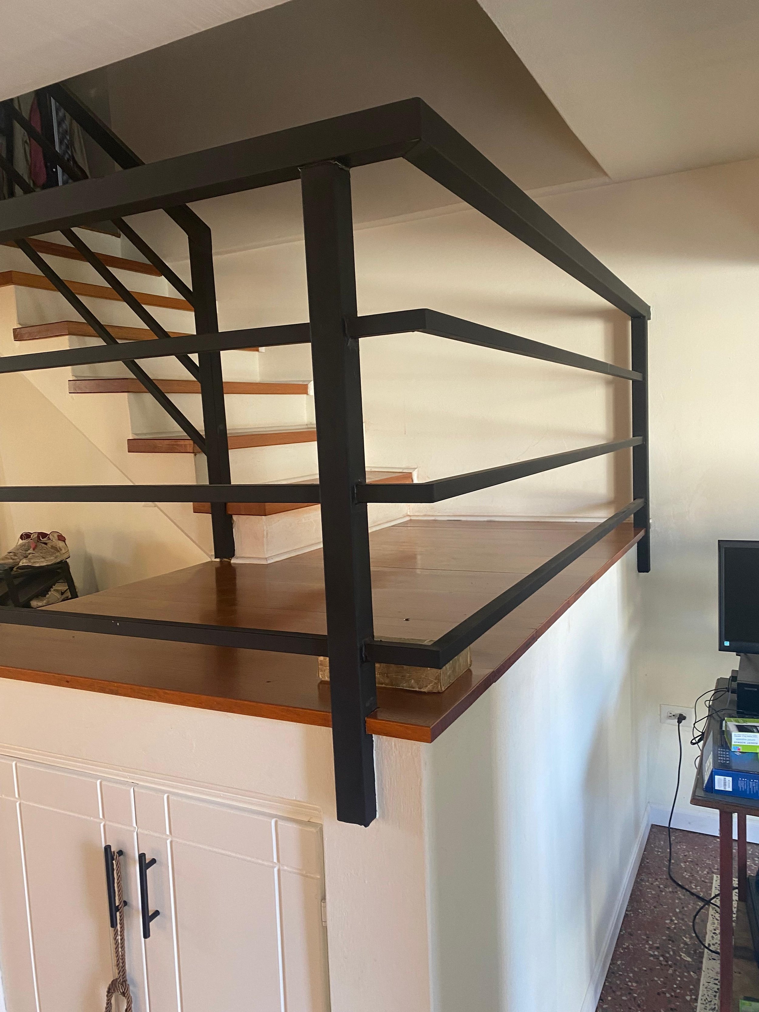 Interior view of a house showing a staircase with wooden steps and a black metal railing. The staircase is against a white wall, and a television and some electronics are visible on a table below.