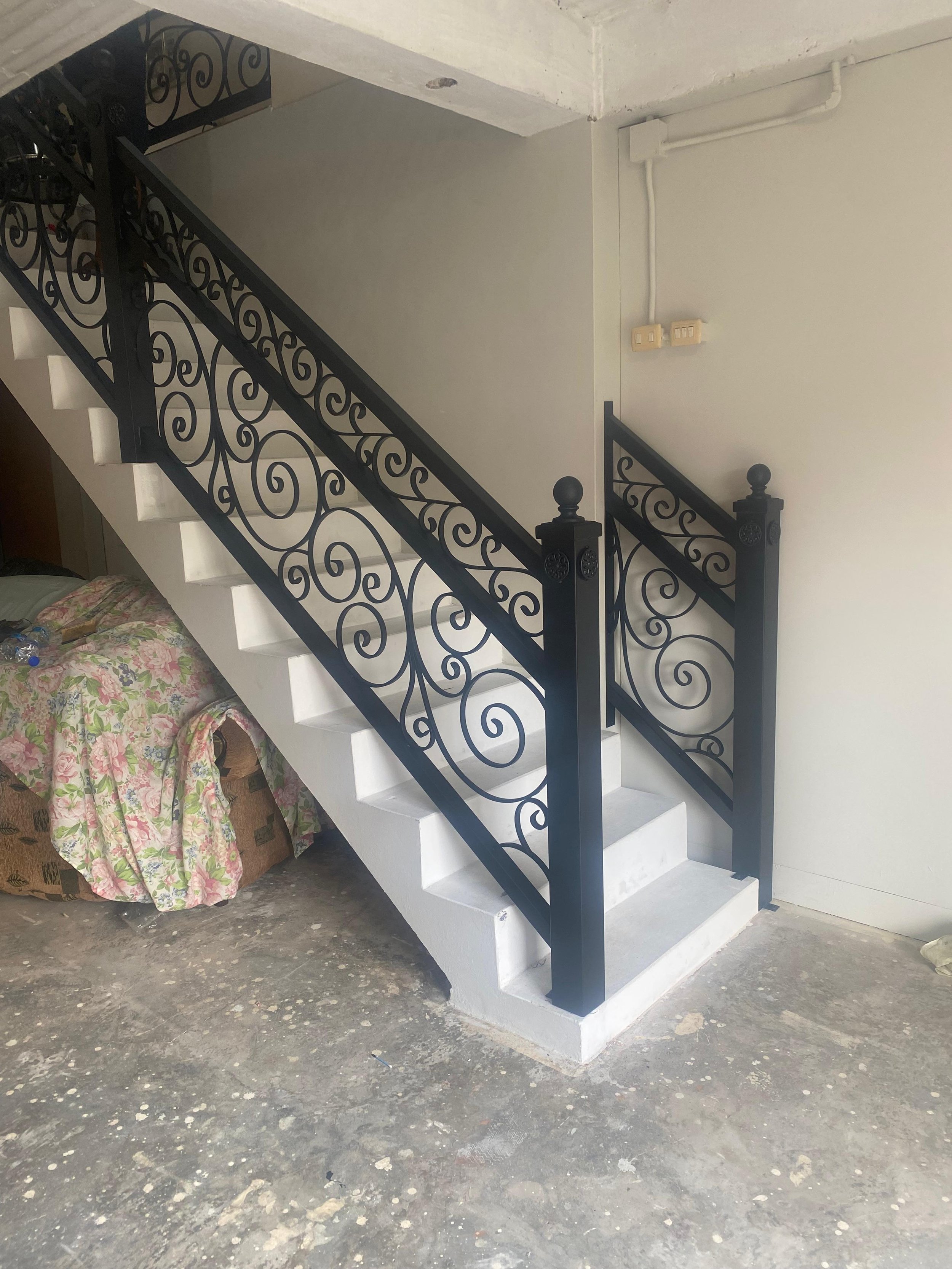 A staircase with black decorative wrought iron railings featuring swirling patterns, set against a plain white wall with visible electrical switches and wiring. The staircase has white steps, and there is furniture, including a covered piece with flo