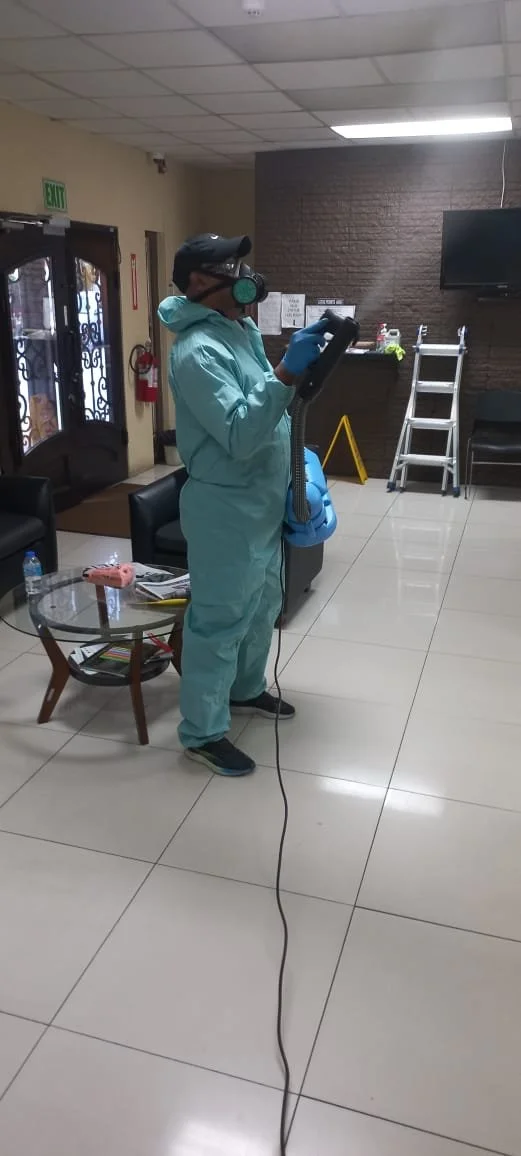 A person wearing a full personal protective suit, gloves, and a mask with a filter is disinfecting or cleaning a room with a sprayer in their hand.