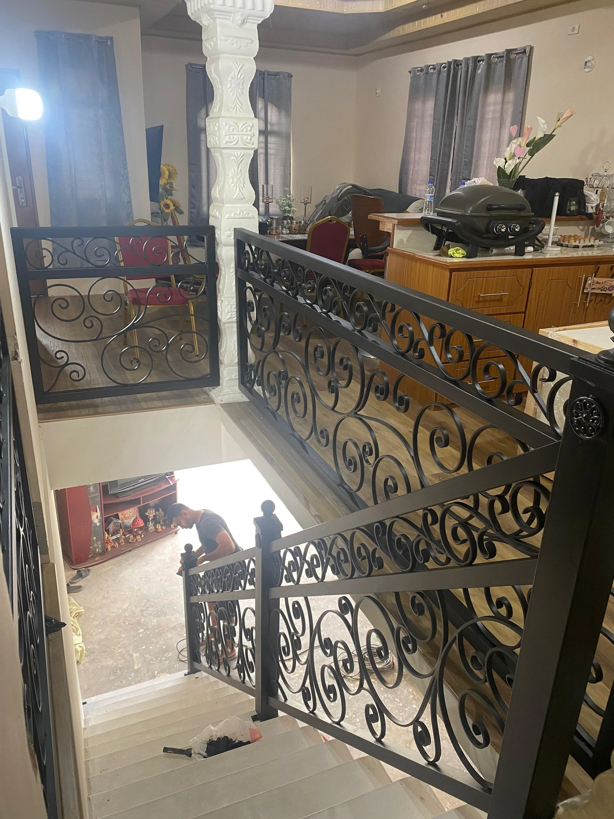 View from the top of a staircase looking down into a living room and kitchen area. A young person is sitting on the floor in the living room, which has a display shelf with decorations. The kitchen counter has a grill, water bottles, and various item