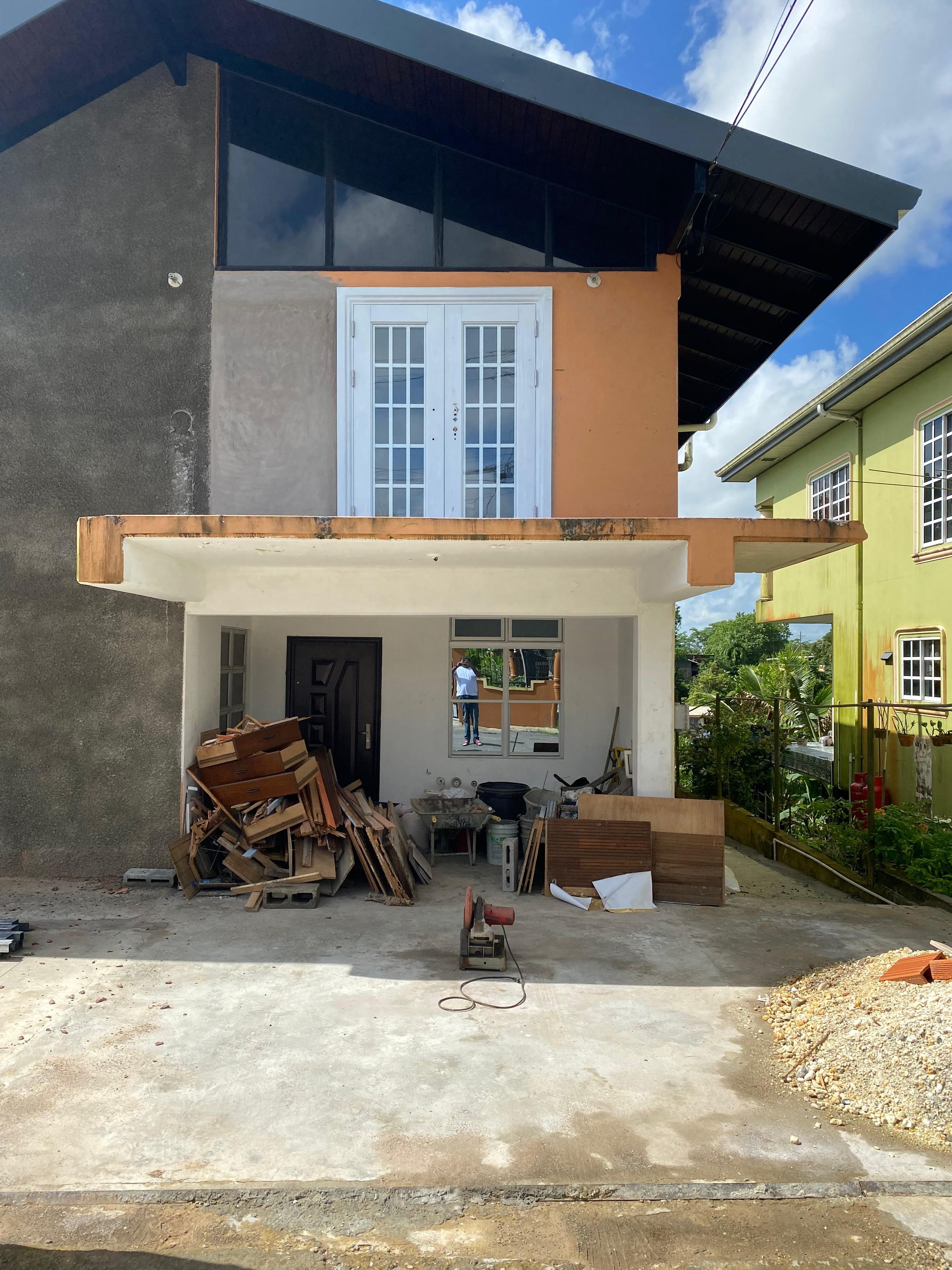 Construction site of a modern house with an unfinished facade, construction materials, and machinery in the foreground, and neighboring houses in the background.