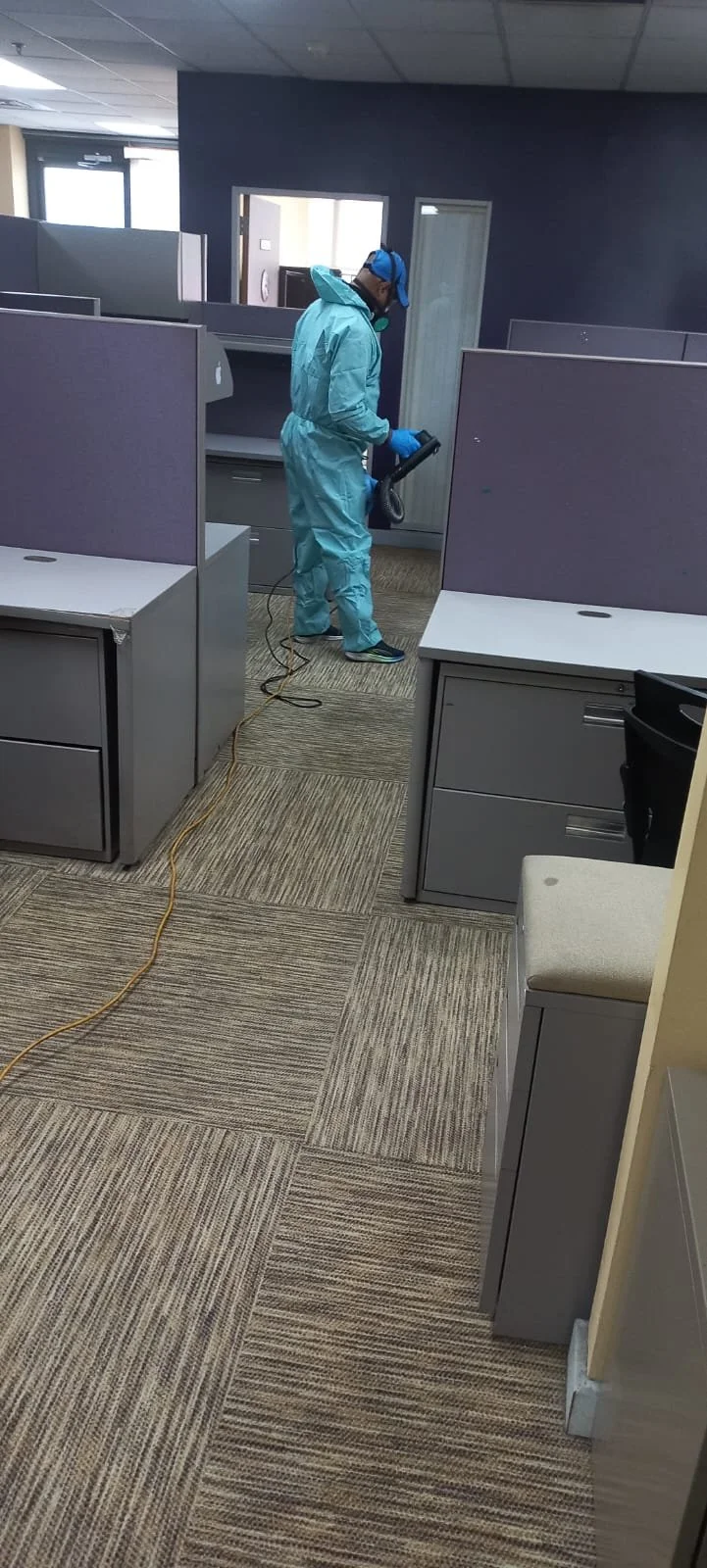 A man in a blue protective suit, gloves, cap, and mask disinfects an office cubicle with a handheld sprayer in a partially empty office space.