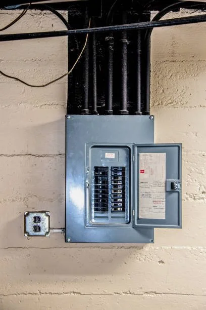 Electrical panel with open door on a beige wall, connected to multiple black cables overhead, with a wall outlet nearby.