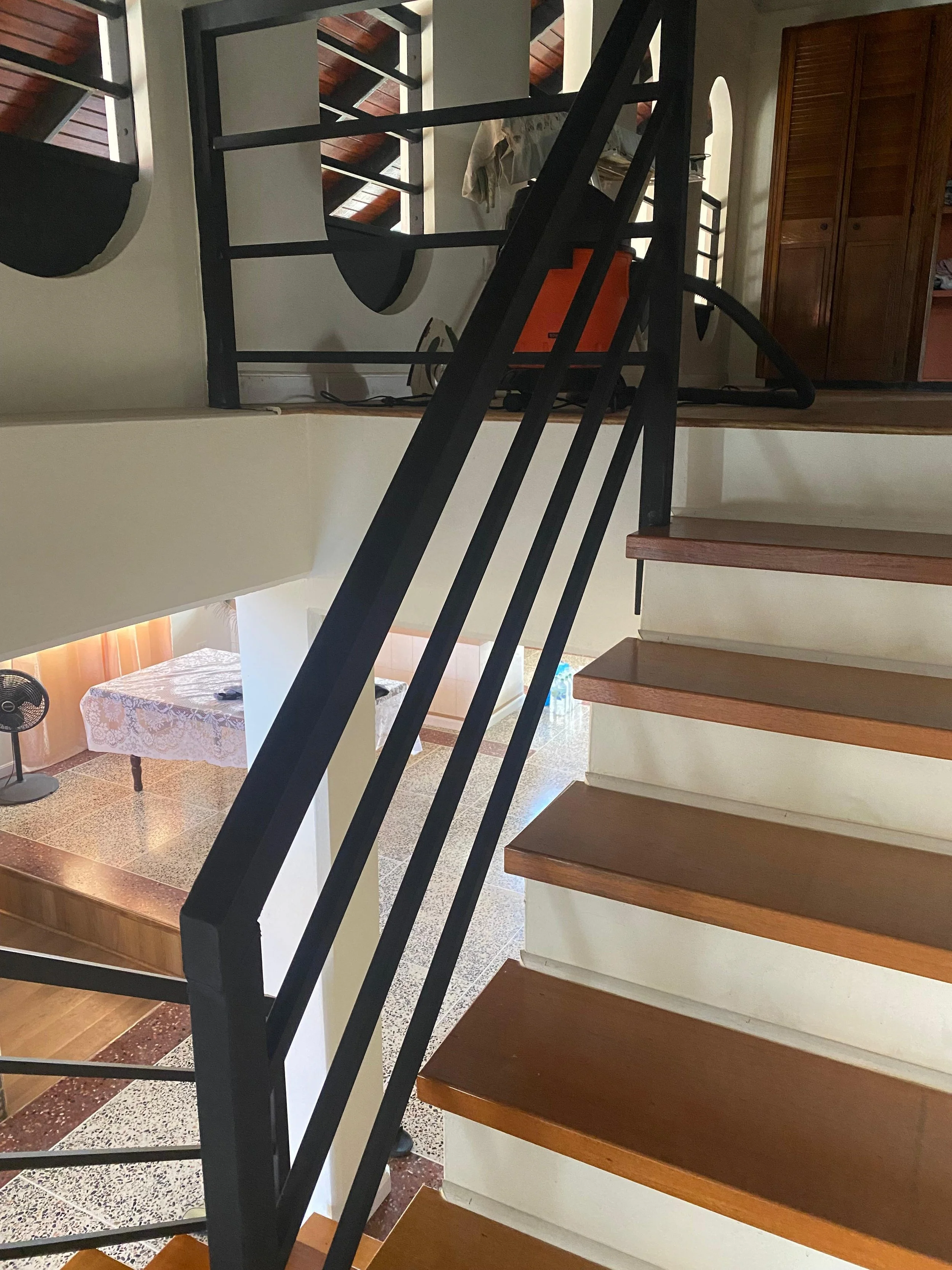 Interior view of a staircase leading to an upper floor, with wooden steps and black metal railing, overlooking a tiled lower level with a table, a fan, and a partly visible wooden wardrobe.