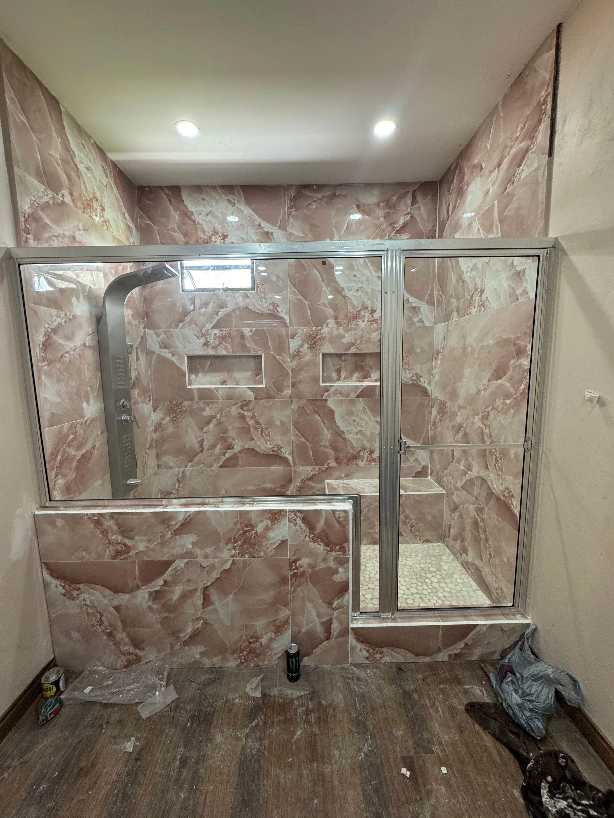Bathroom shower with pink marble tiles, glass door, shower head, and miscellaneous construction materials on the floor.