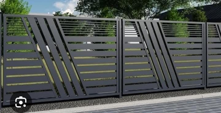Modern metal fence with diagonal slats, painted dark gray, installed on a sidewalk with grass and trees in the background.