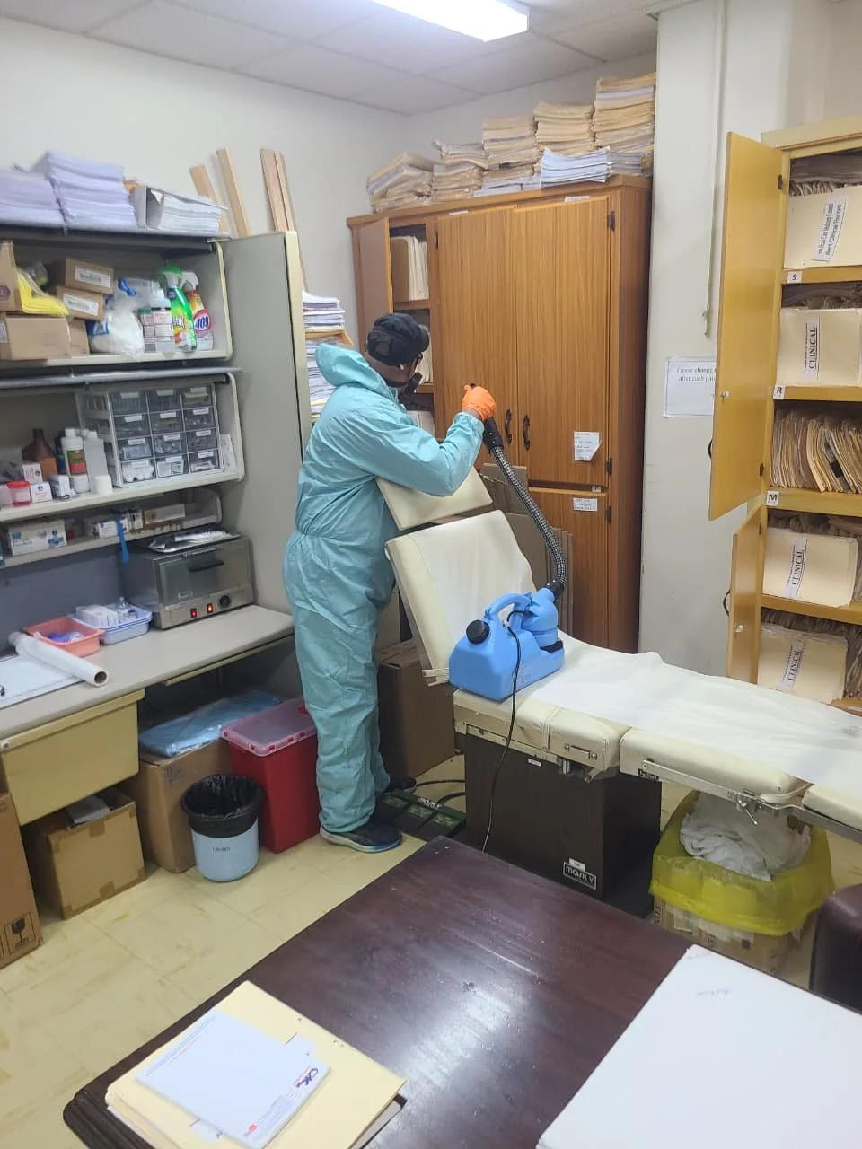 A person in medical scrubs and gloves cleaning an office space with a vacuum-like device; surrounded by shelves of papers, files, and supplies.