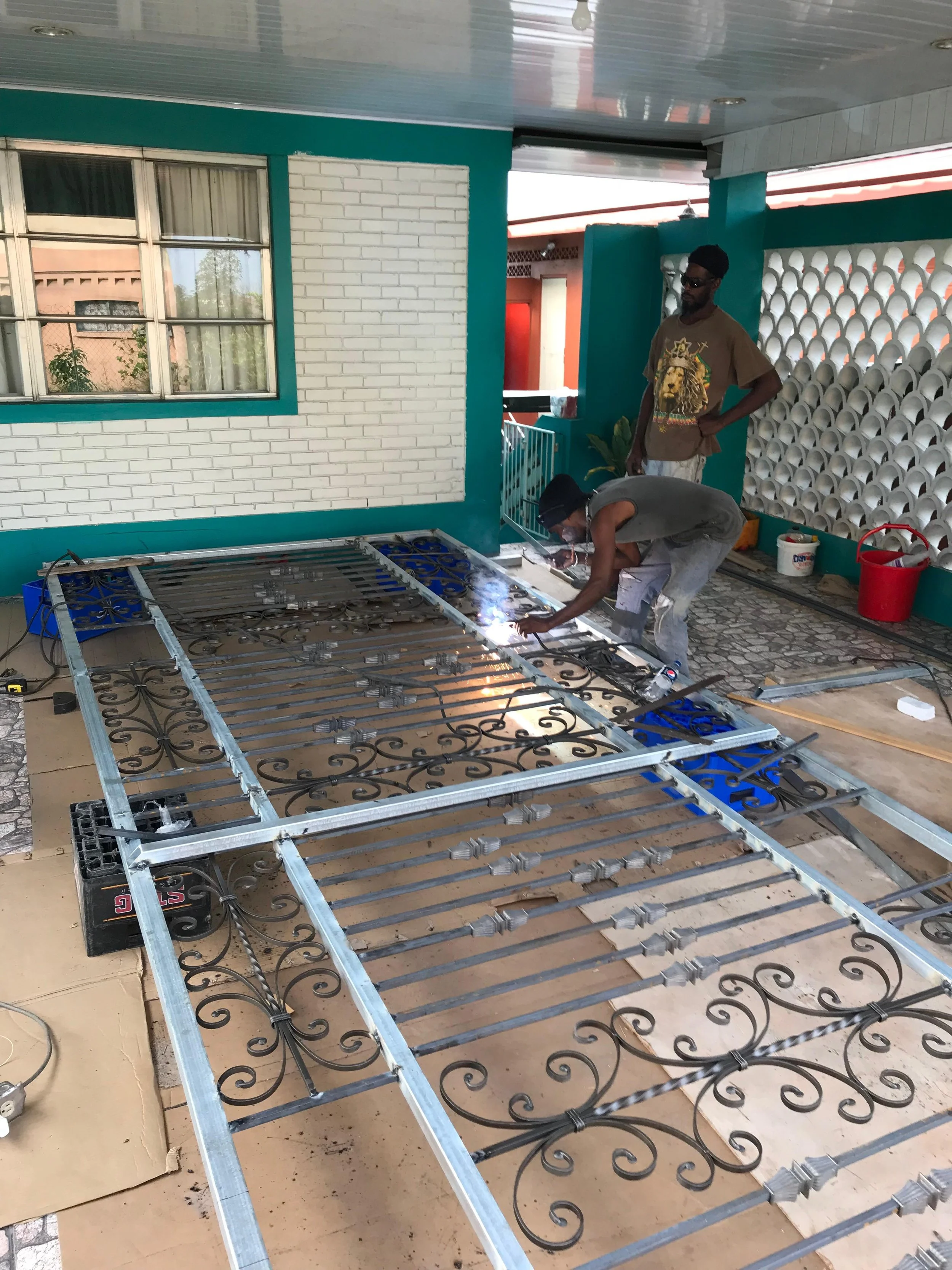 Two men are working on installing wrought iron balcony railings outside a building. One man is kneeling, welding or fastening the metal, while the other is standing nearby, observing. The building has teal frames around white brick walls and patterne