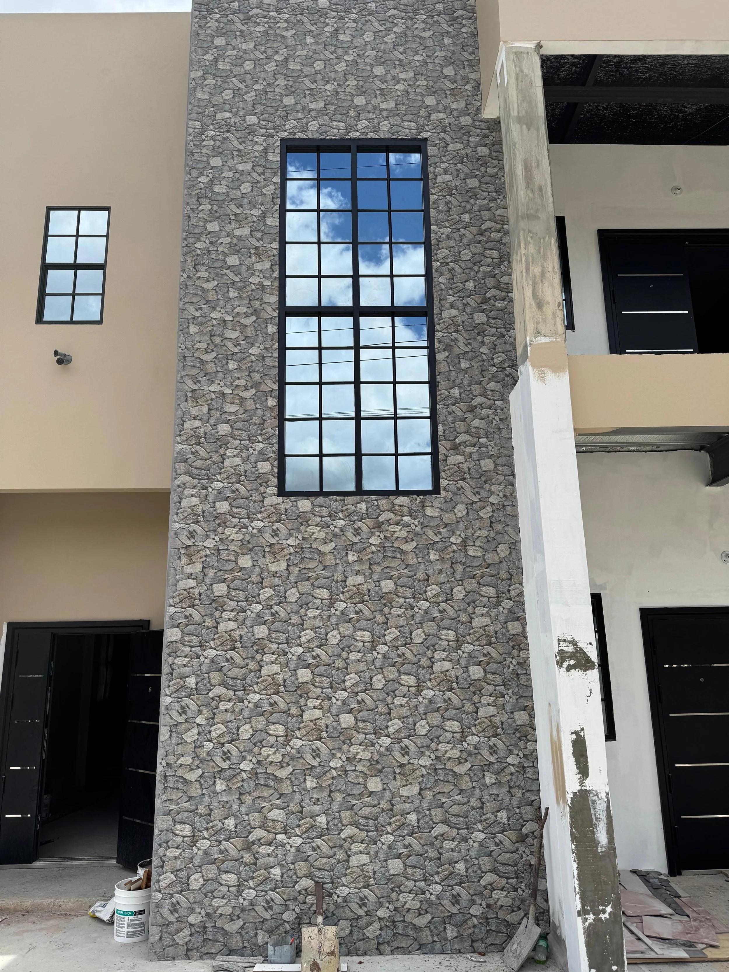Exterior view of a modern building under construction, featuring a textured stone wall, large window reflecting the sky, and black doors.