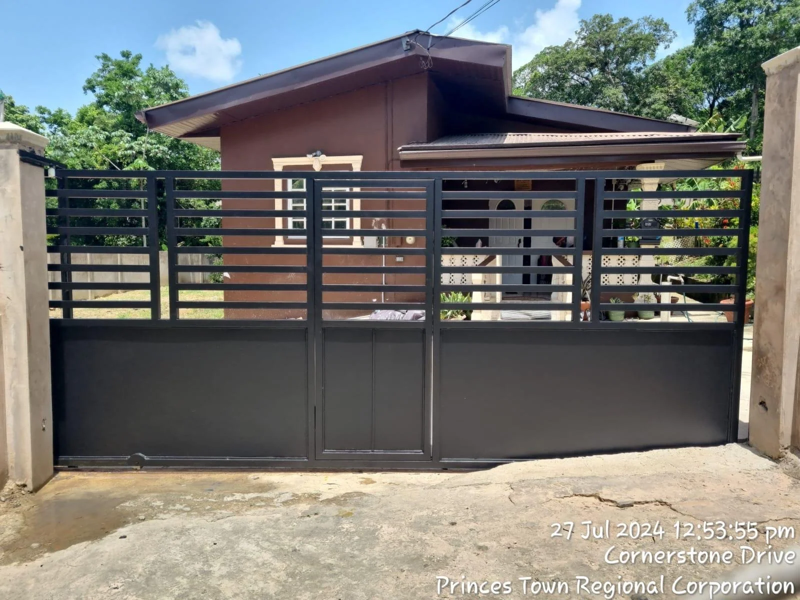 A black metal gate with horizontal slats and a smaller pedestrian door, situated at the entrance of a house with a brown exterior and white trim. Green trees and a partially visible house are in the background. The sky is blue with scattered clouds. 