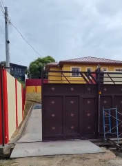 A house with a yellow upper section and a dark brown gate in front. The house has a red roof and a concrete driveway, with a sidewalk and some trees in the background under a cloudy sky.