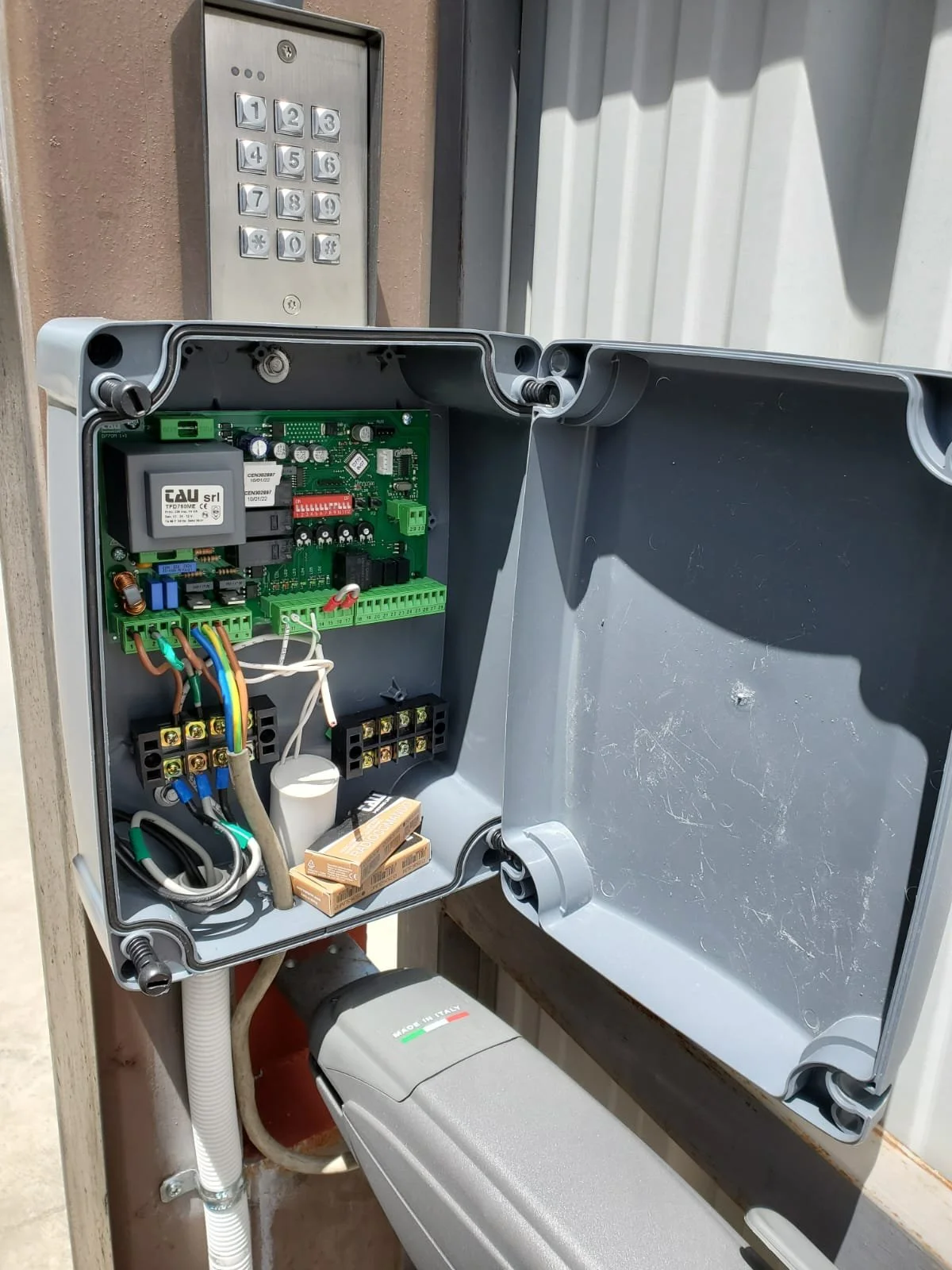 Open outdoor electrical control box with circuit board, wiring, and components, mounted on a wall near a keypad entry system.