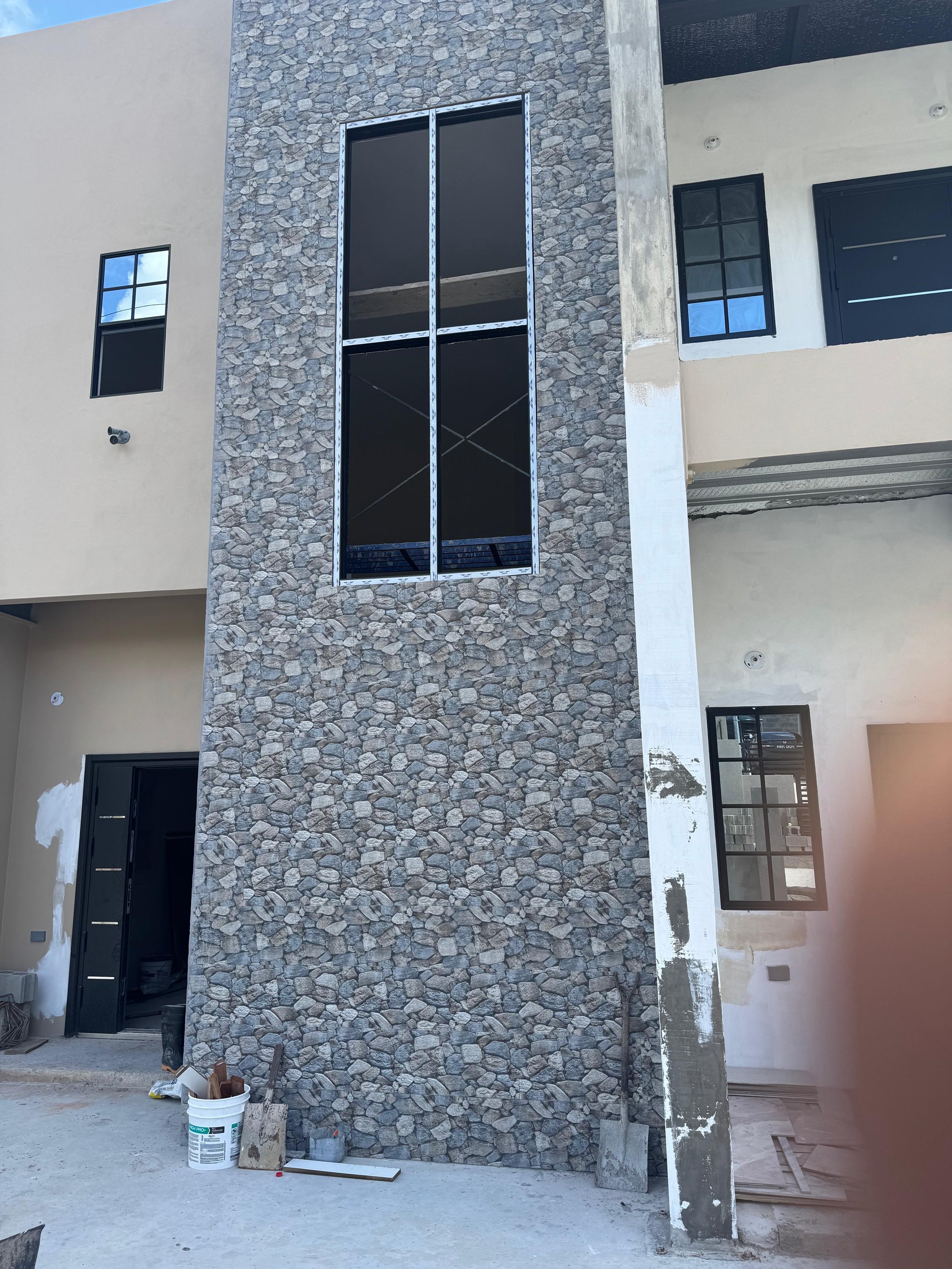 Construction site of a modern building facade with a stone wall and black framed windows, some construction materials and tools visible on the ground.