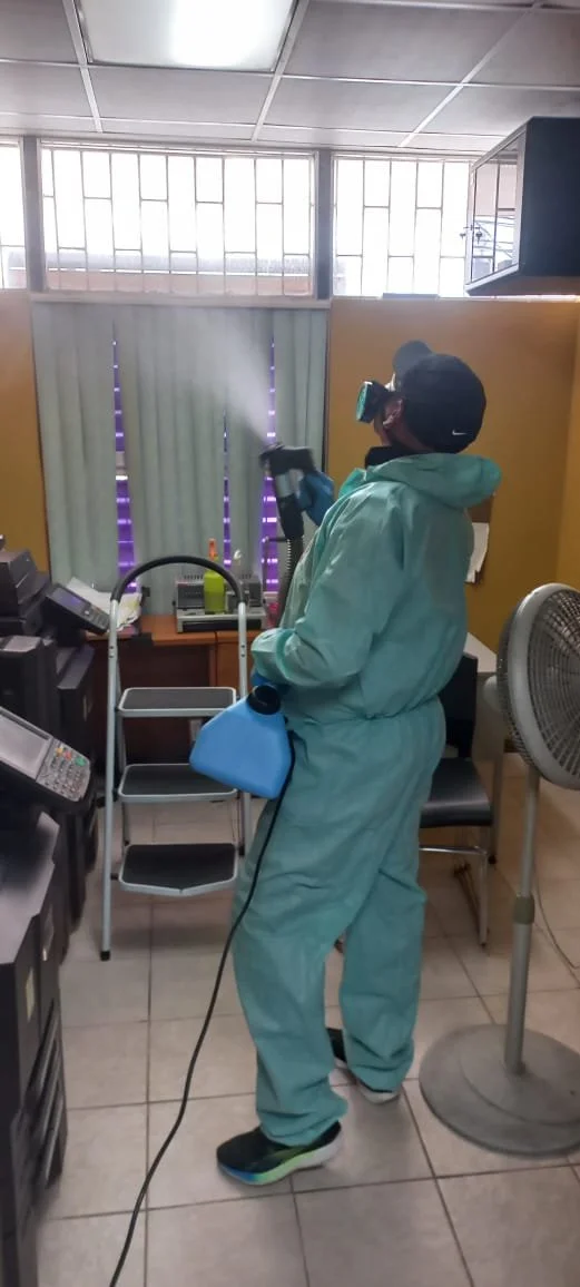 Person in full protective suit disinfecting an office area with cleaning equipment, including a spray wand and a blue cleaning cloth.