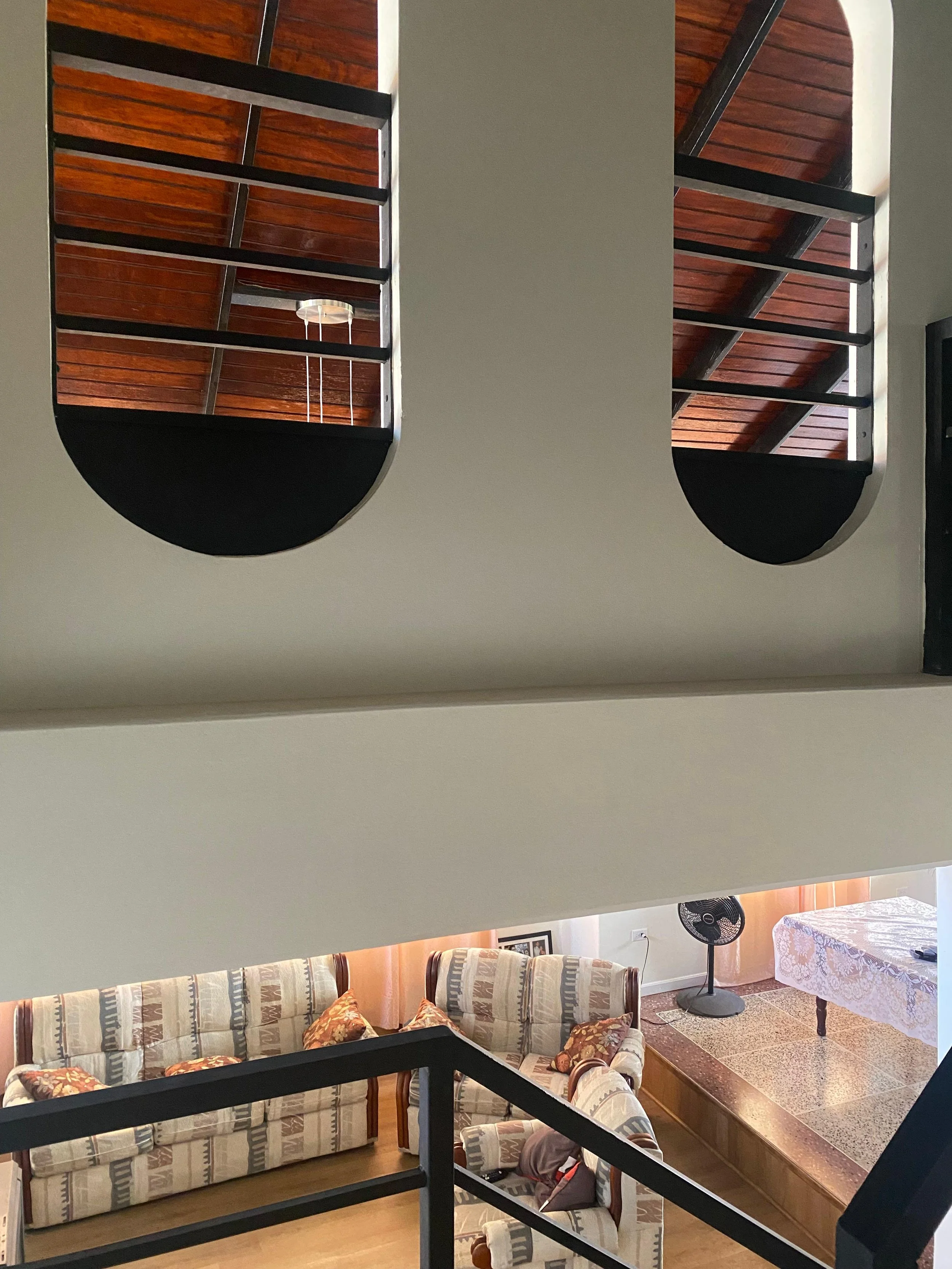 Living room staircase with two wooden risers and black metal railing, viewed from below looking up, with a patterned sofa, table with lace tablecloth, fan, and framed picture on the wall in the background.