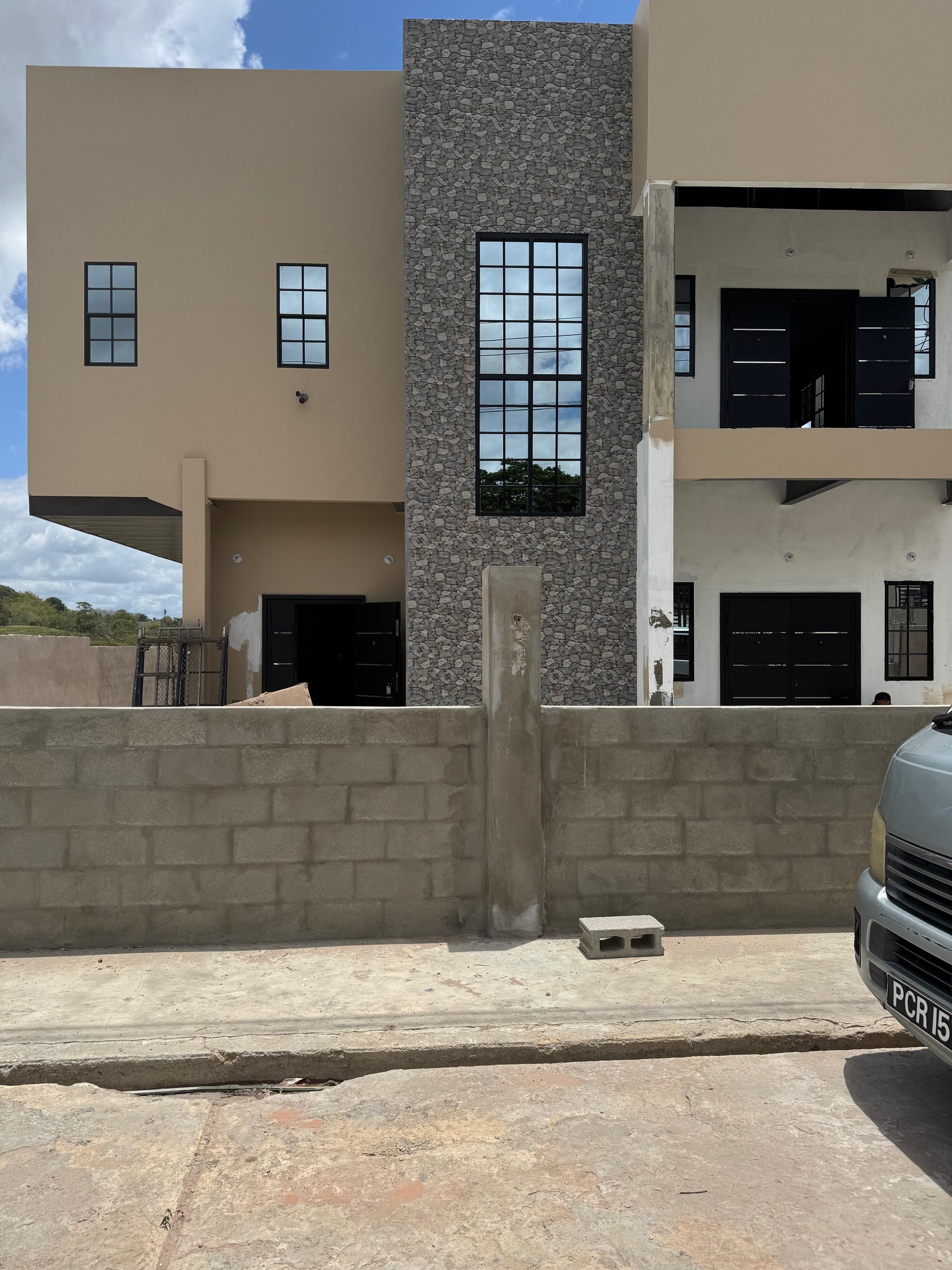 The image shows a modern, multi-story building under construction or renovation, with a beige and stone exterior. A concrete block wall and a portion of a parked vehicle are visible in the foreground, with a concrete road surface and a low wall separ
