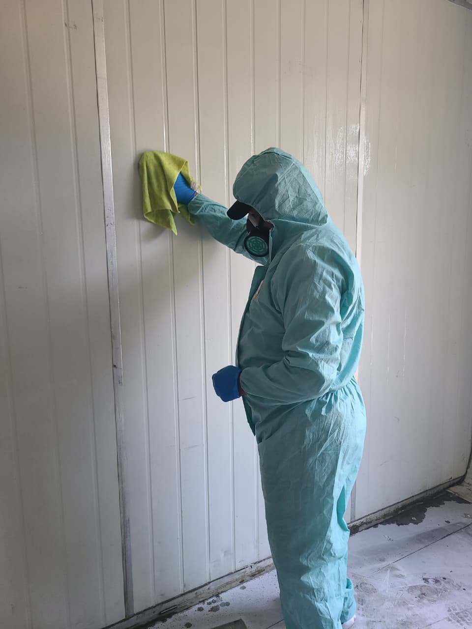 A person dressed in light blue protective clothing, gloves, and a mask cleaning a wall with a yellow cloth.