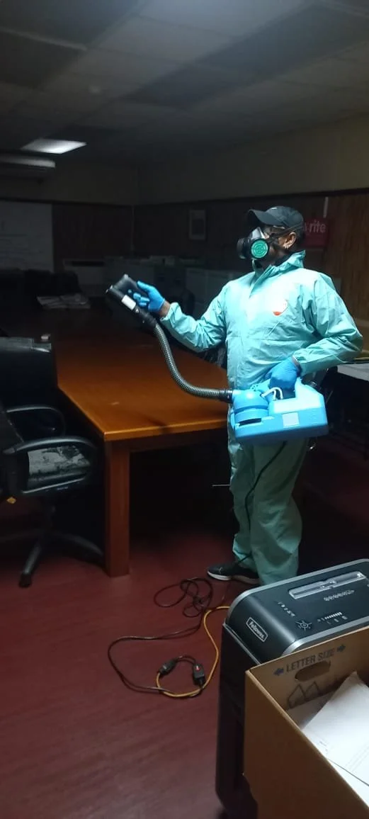 A person wearing a protective suit, gloves, a face mask, and a cap, holding cleaning equipment, in a dark office room.
