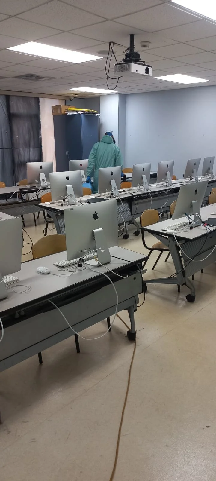 Empty classroom with multiple Apple iMac computers on desks, a person in protective gear standing near a cupboard, and a ceiling-mounted projector.