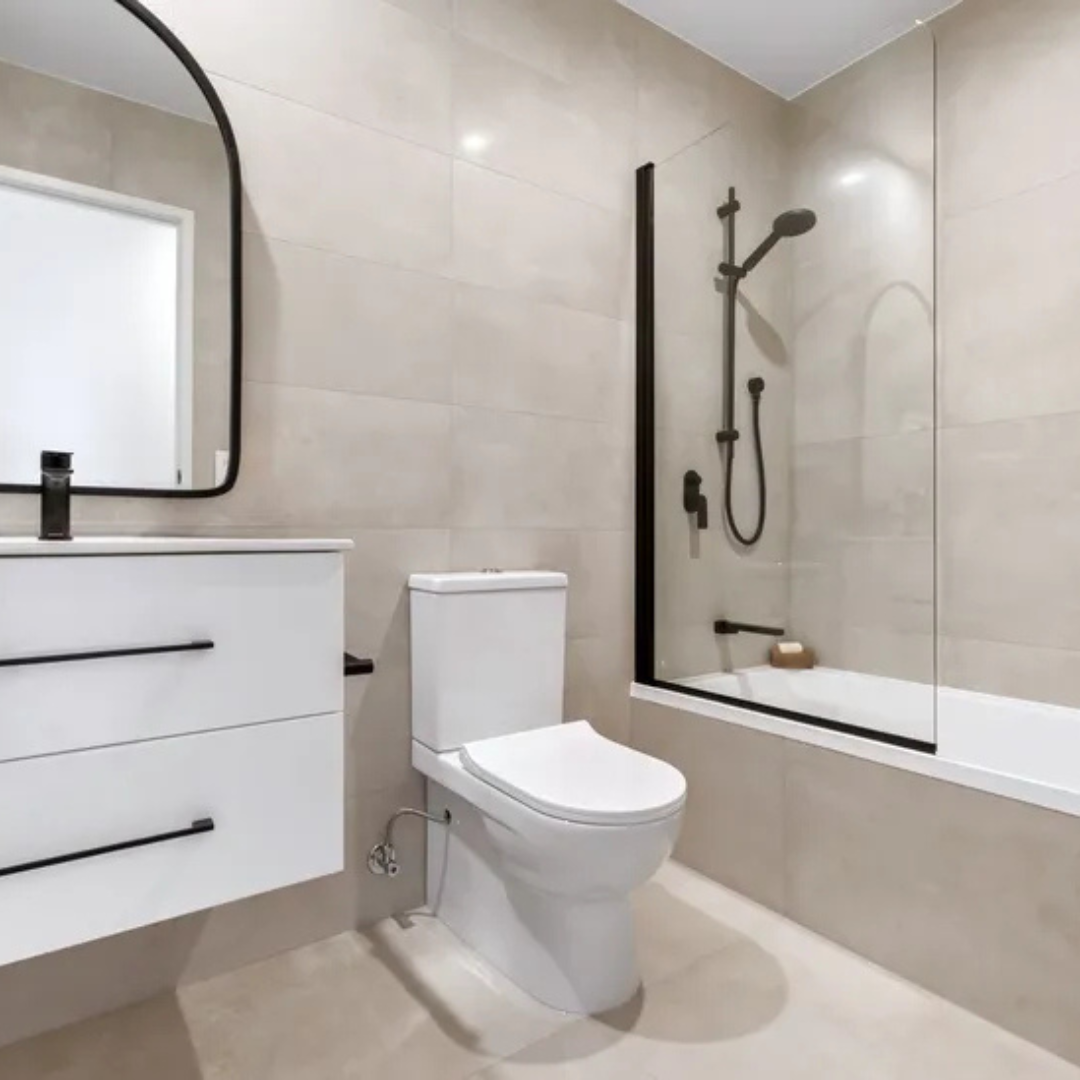 Modern bathroom with beige tiled walls, a white toilet, a floating white vanity with black handles, a black framed mirror, and a walk-in shower with a glass partition and black fixtures.