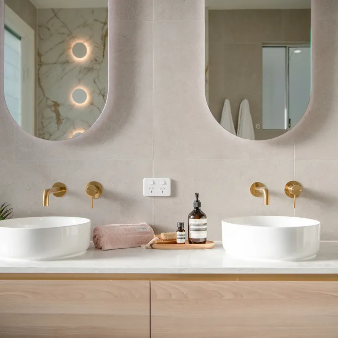 Modern bathroom with double vanity featuring round white vessel sinks, gold wall-mounted faucets, two mirrors, a pink towel, and bathroom toiletries on a tray.