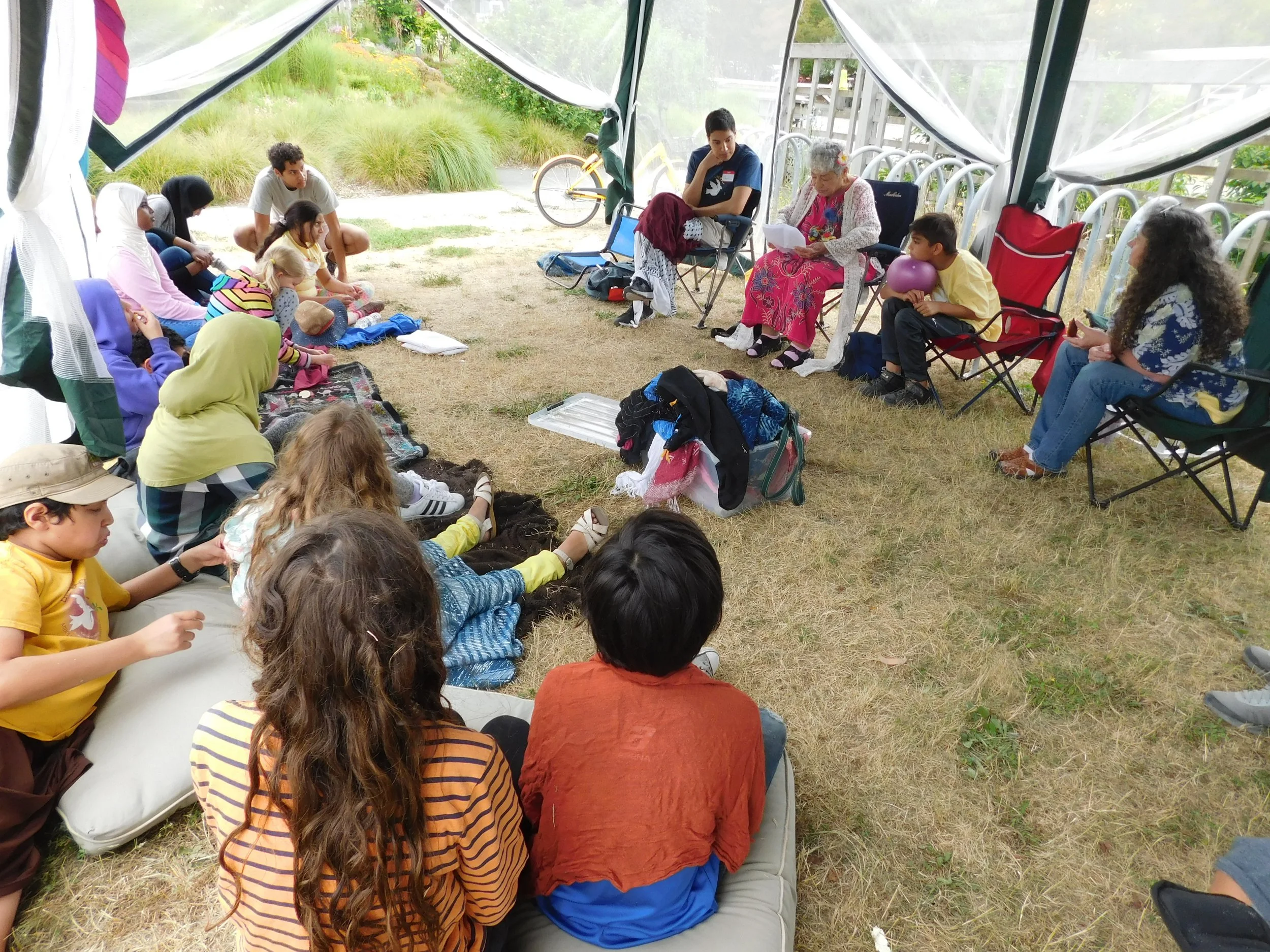A group of people sitting and kneeling in a semi-circle under a tent, engaged in a gathering or discussion outdoors. Some are sitting on blankets and chairs, with greenery and a bicycle visible outside.