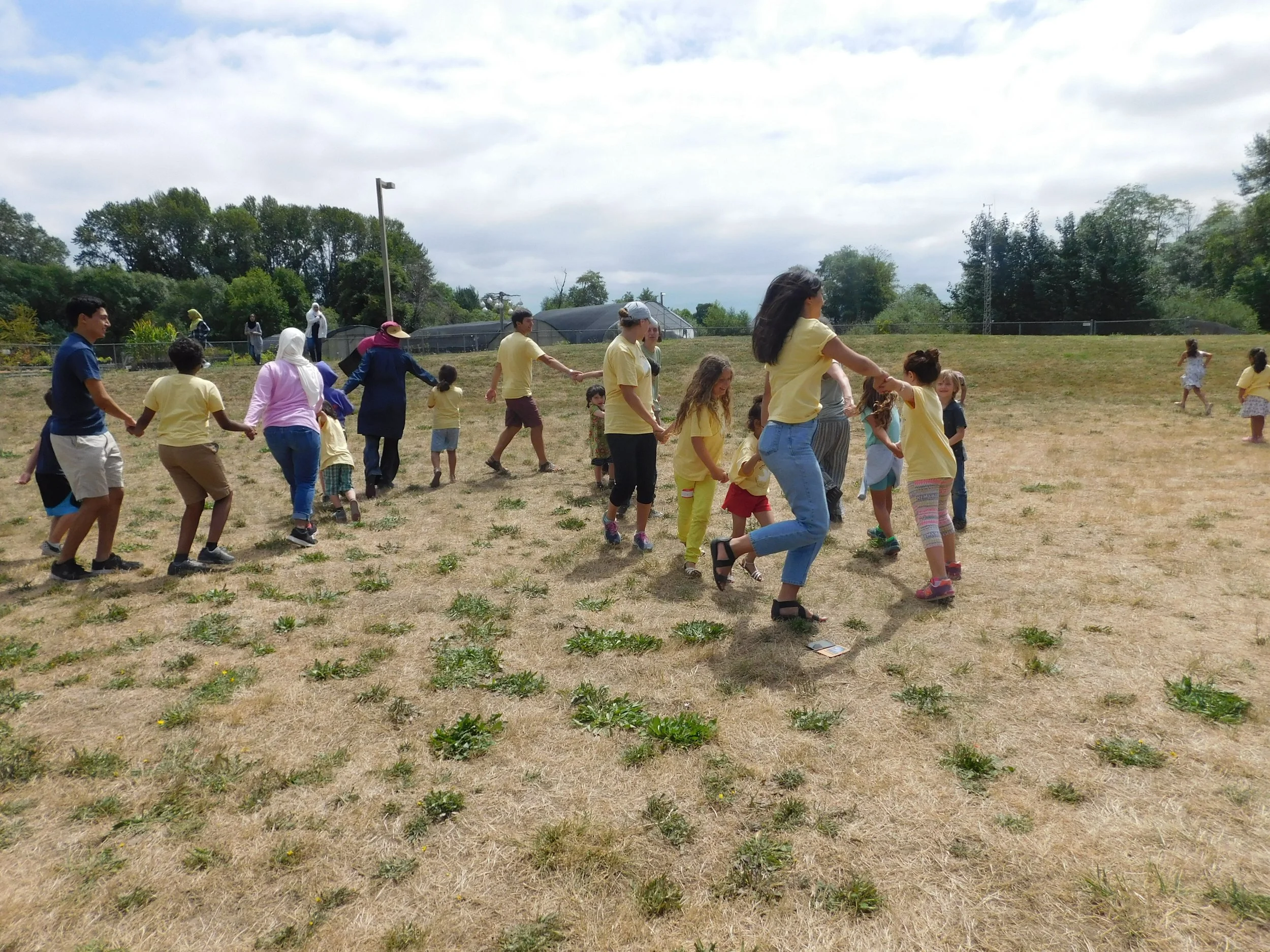 Children and adults holding hands and dancing in a circle outdoors on a grassy field under cloudy sky.
